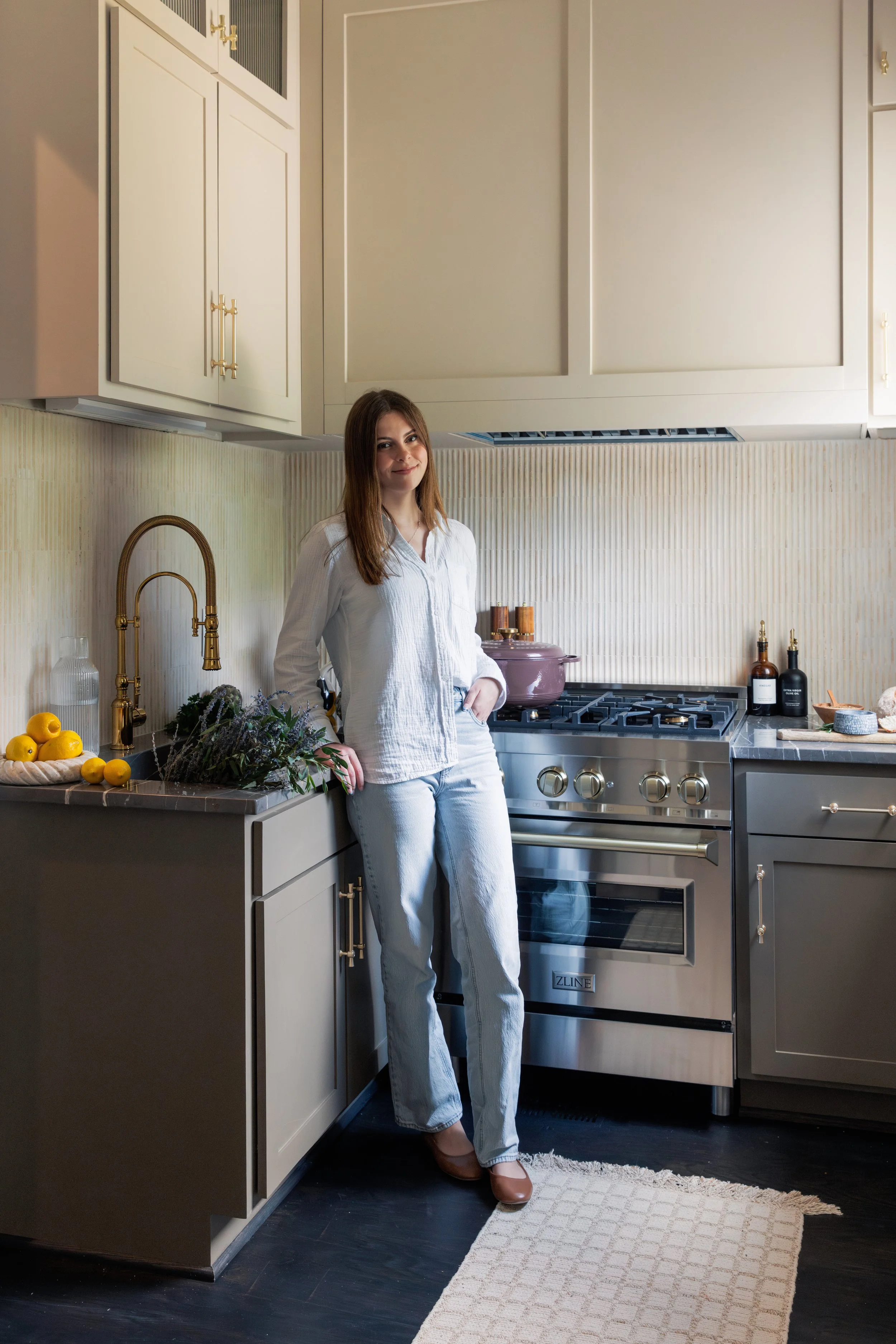 A woman in a white shirt and light blue jeans stands in a modern kitchen, leaning against the counter near a stove, with lemons, herbs, and kitchen utensils on the counter.