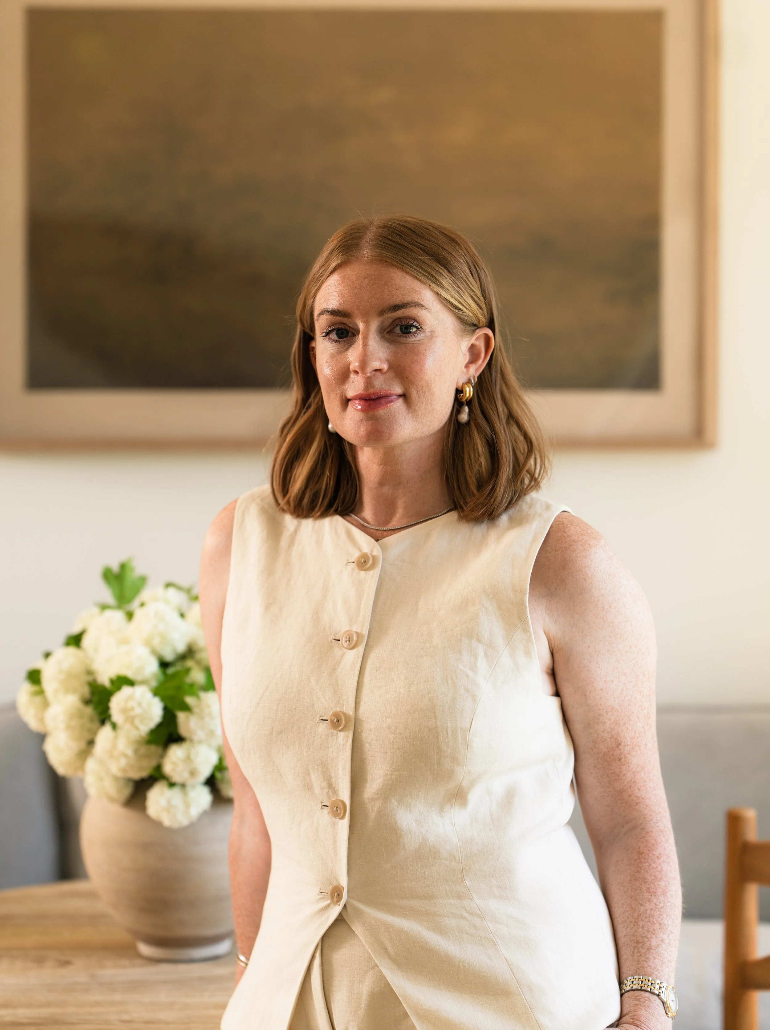 A woman with shoulder-length reddish hair, wearing a cream-colored sleeveless dress with buttons, standing indoors near a table with a bouquet of white flowers in a vase, and looking at the camera with a slight smile.