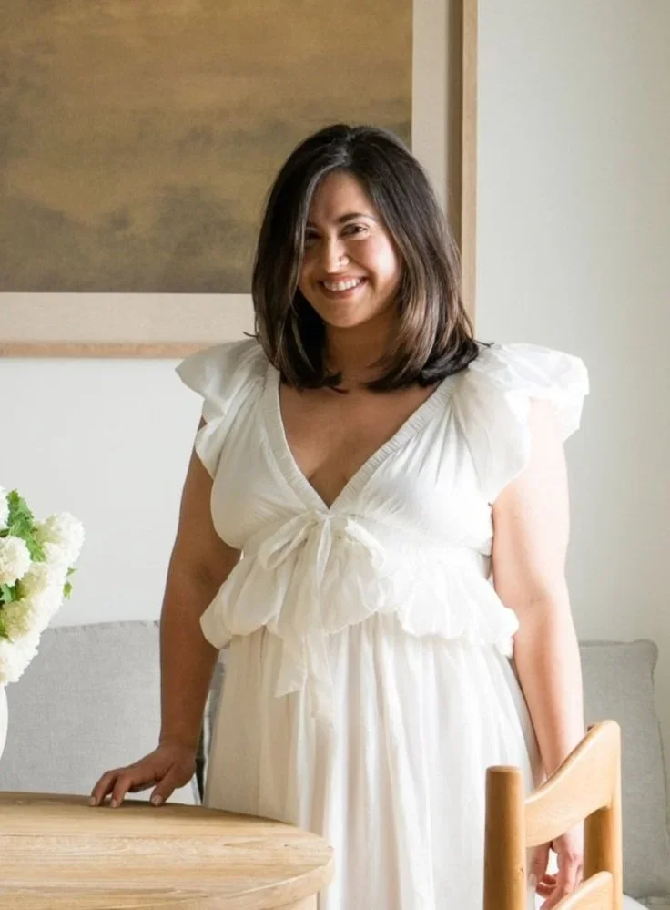 A woman with shoulder-length dark hair wearing a white dress with puffed sleeves, standing indoors near a table with white flowers and a wooden chair, smiling at the camera.