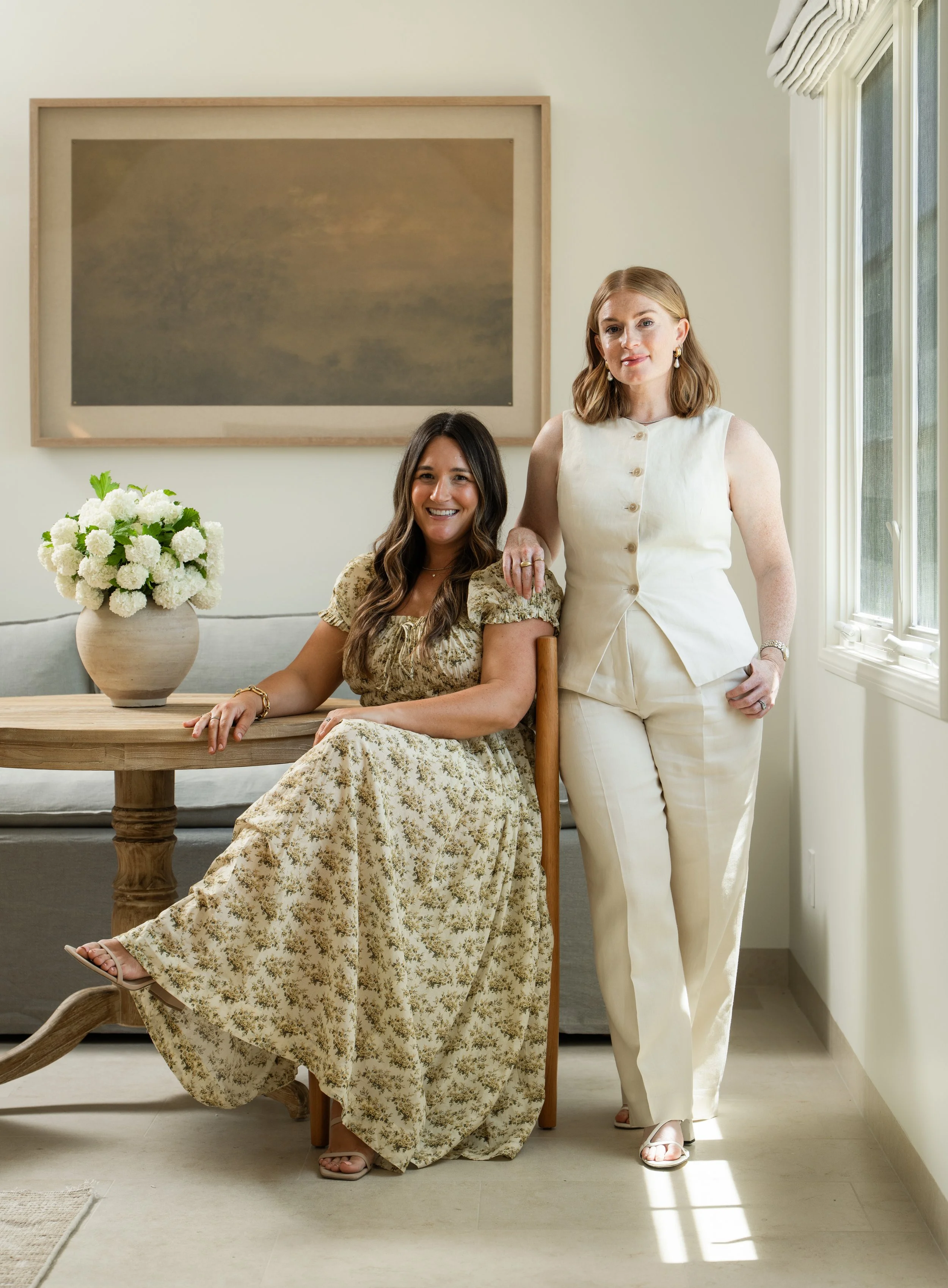 Two women are posing in a well-lit living room near a window. One woman is seated at a wooden table with a large white floral arrangement, and the other woman is standing beside her with her hand resting on her shoulder. Both are smiling.