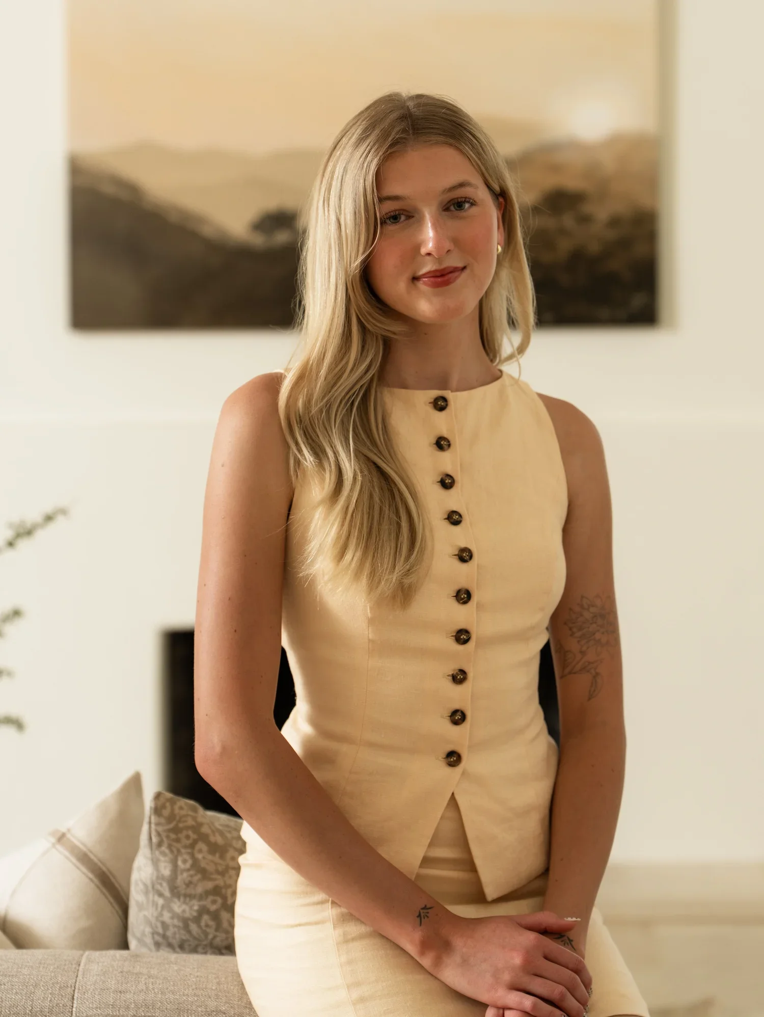 Young woman with long blond hair and tattoos, wearing a cream-colored sleeveless dress, sitting on a beige sofa in a living room with a landscape painting in the background.