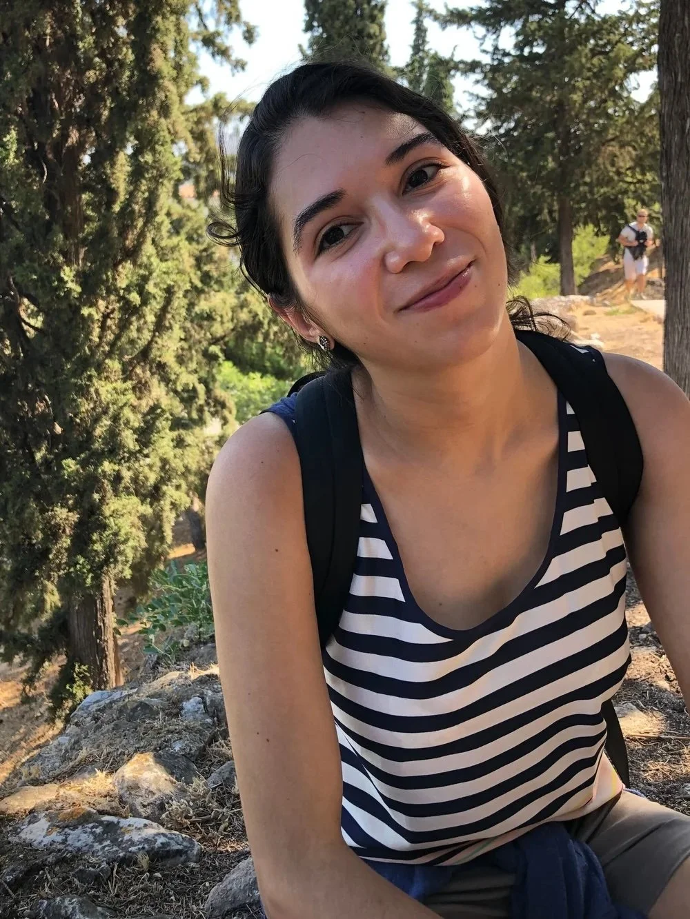 A young woman with dark hair smiling outdoors in a wooded area, wearing a striped tank top and carrying a backpack.