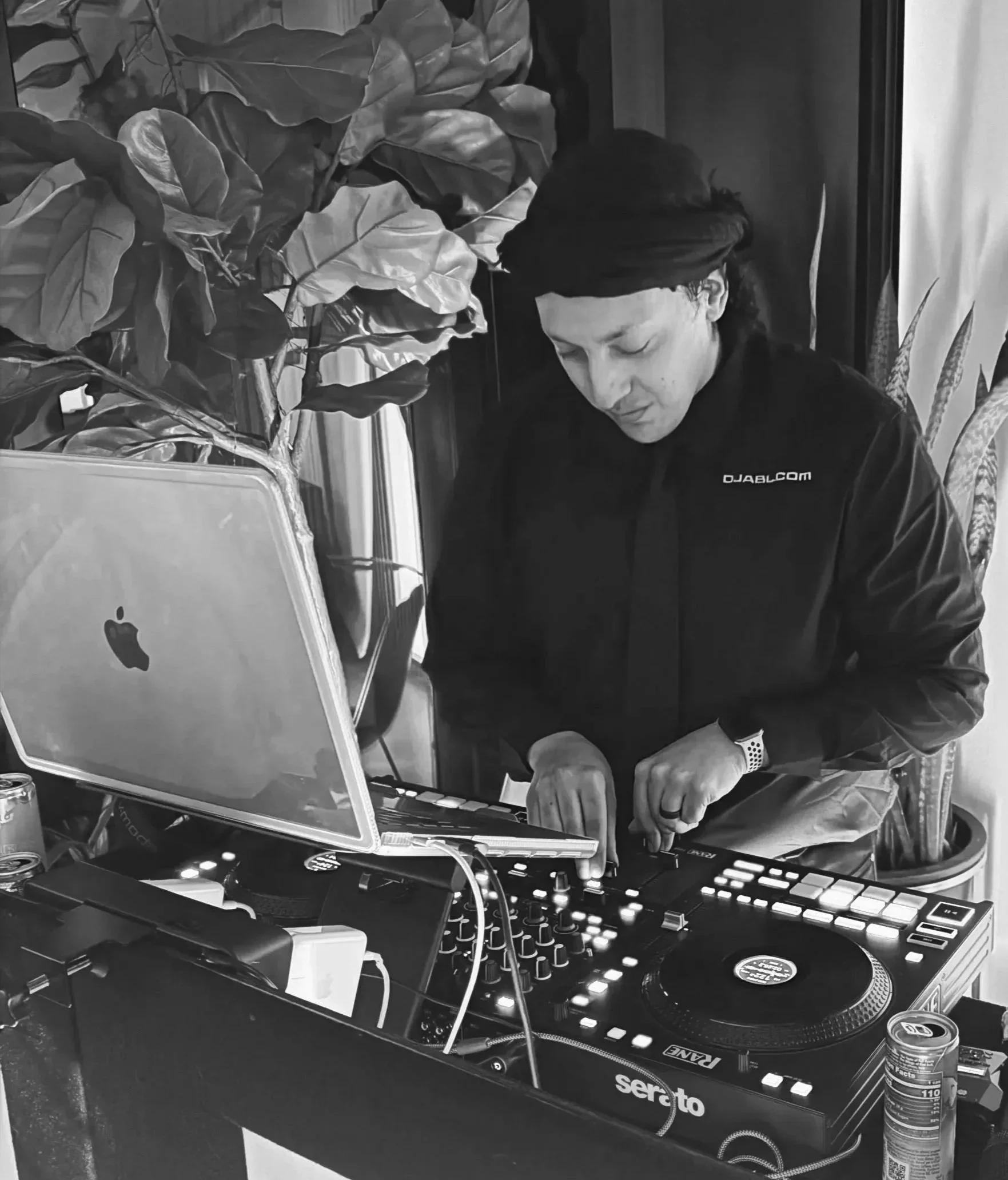 A person wearing a dark hat and shirt working with DJ equipment, including a Pioneer Serato DJ controller, turntable, a laptop with an Apple logo, with a plant partially visible in the background.