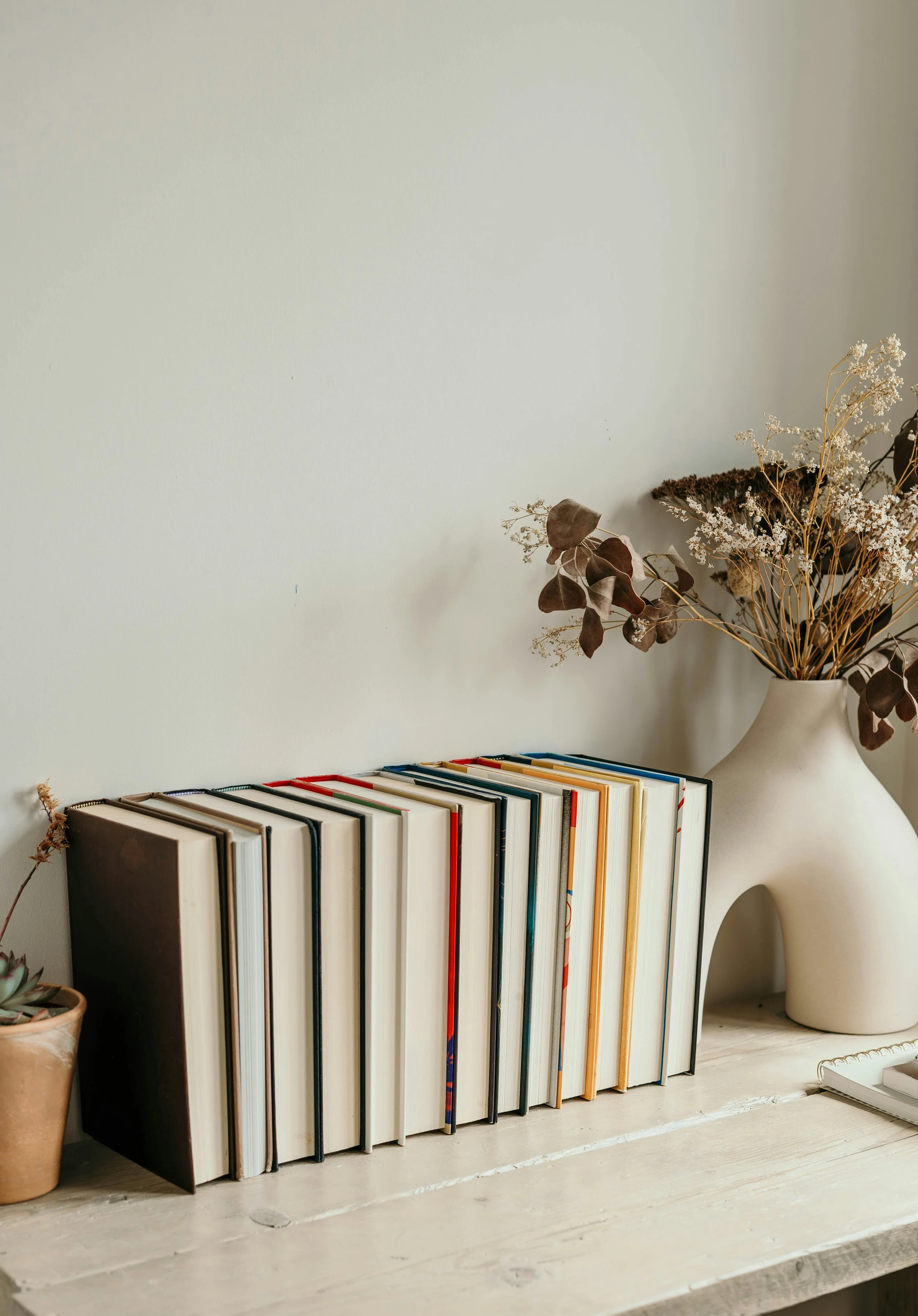A row of books and notebooks on a white wooden shelf, decorative dried flowers in a large beige vase, small potted succulent plant, and a closed notebook to the right.