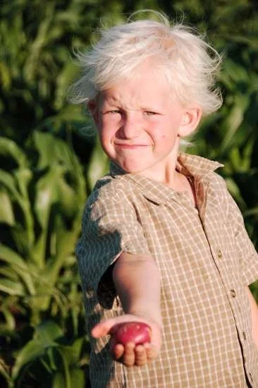 A young girl with messy white hair holding a pink fruit, outdoors in a garden with green plants.