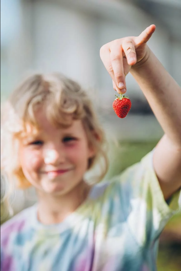 A young girl with curly blonde hair smiling while holding a strawberry up to the camera outdoors.