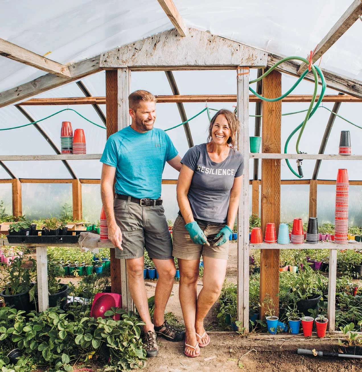 A man and woman smiling inside a greenhouse surrounded by potted plants and gardening supplies, with colorful cups and gardening hoses visible. The woman is wearing a shirt with the word 'RESILIENCE'.