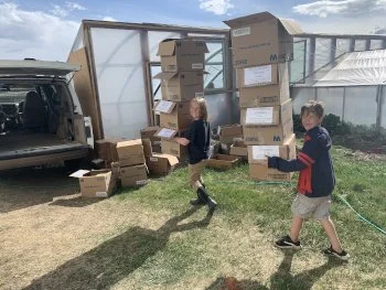 Two children carrying boxes outdoors near a van and a large stack of cardboard boxes.