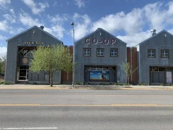 Three industrial-style buildings with a blue sky and clouds in the background, trees in front, and a downtown street with a double yellow line.