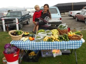 A woman holding a young boy at a farmers market stall with fresh vegetables and fruits on a blue checkered tablecloth.