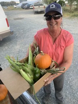 Smiling woman wearing sunglasses and a baseball cap holding a box of fresh vegetables, including corn and a small pumpkin, outdoors with cars in the background.