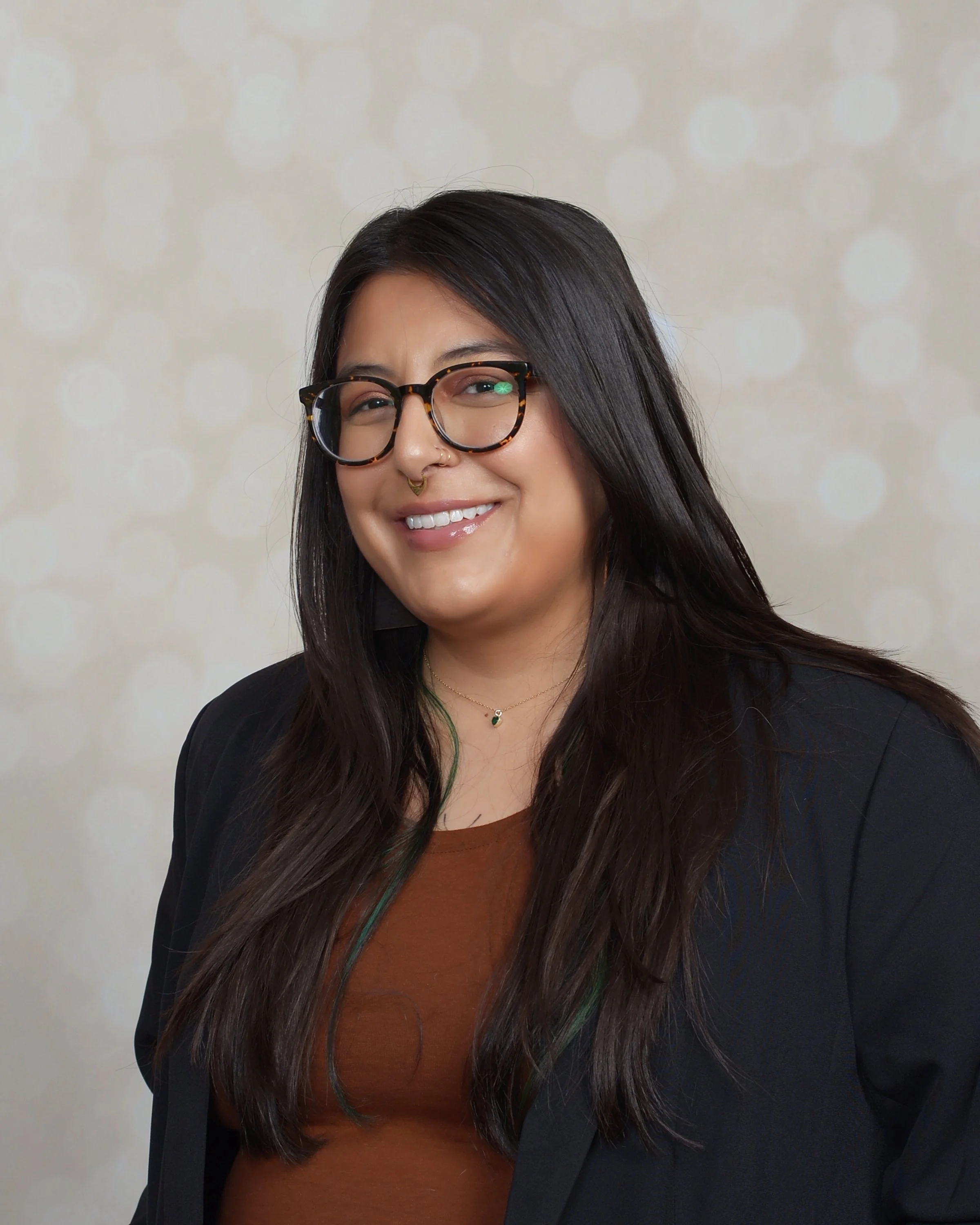 A woman with long dark hair, glasses, and a nose piercing, smiling at the camera, wearing a black blazer and brown top, with a light-colored background.