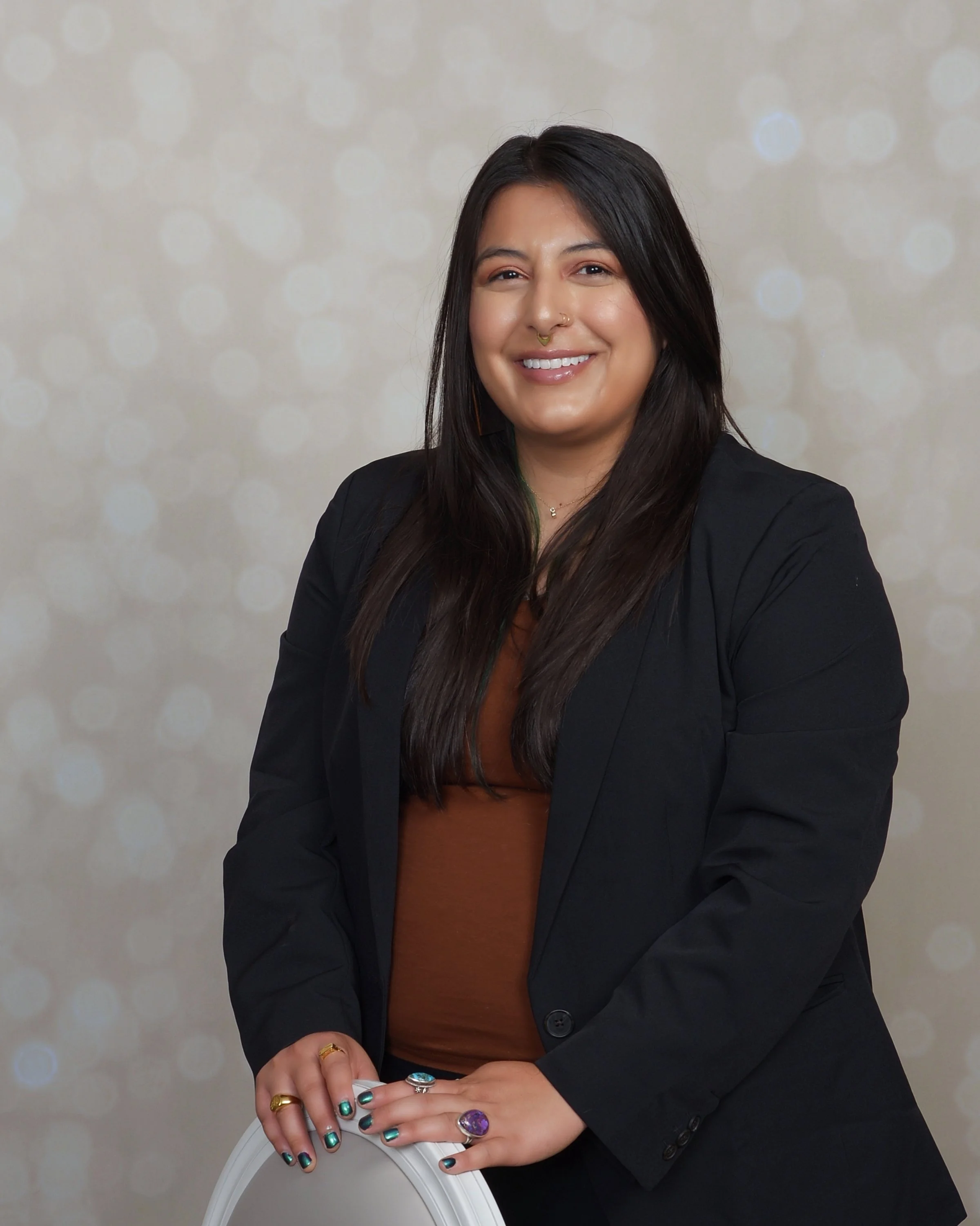 A woman with long dark hair, smiling, wearing a black blazer and multiple rings, seated with her hands on a white chair, against a blurred light-colored background with circular bokeh lights.