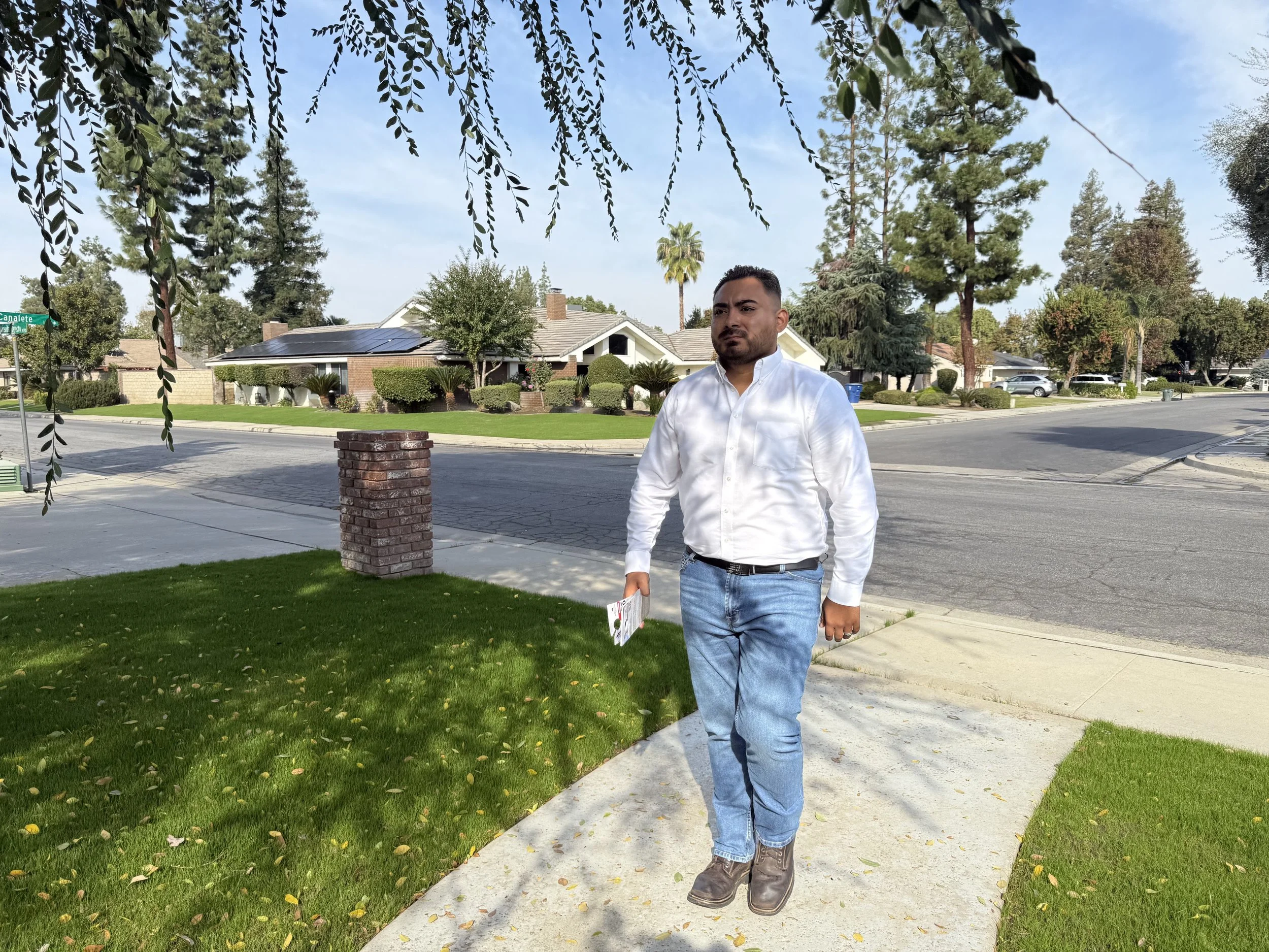 A man in a white shirt and blue jeans walking on a sidewalk in a residential neighborhood, holding some papers, with greenery and houses in the background.