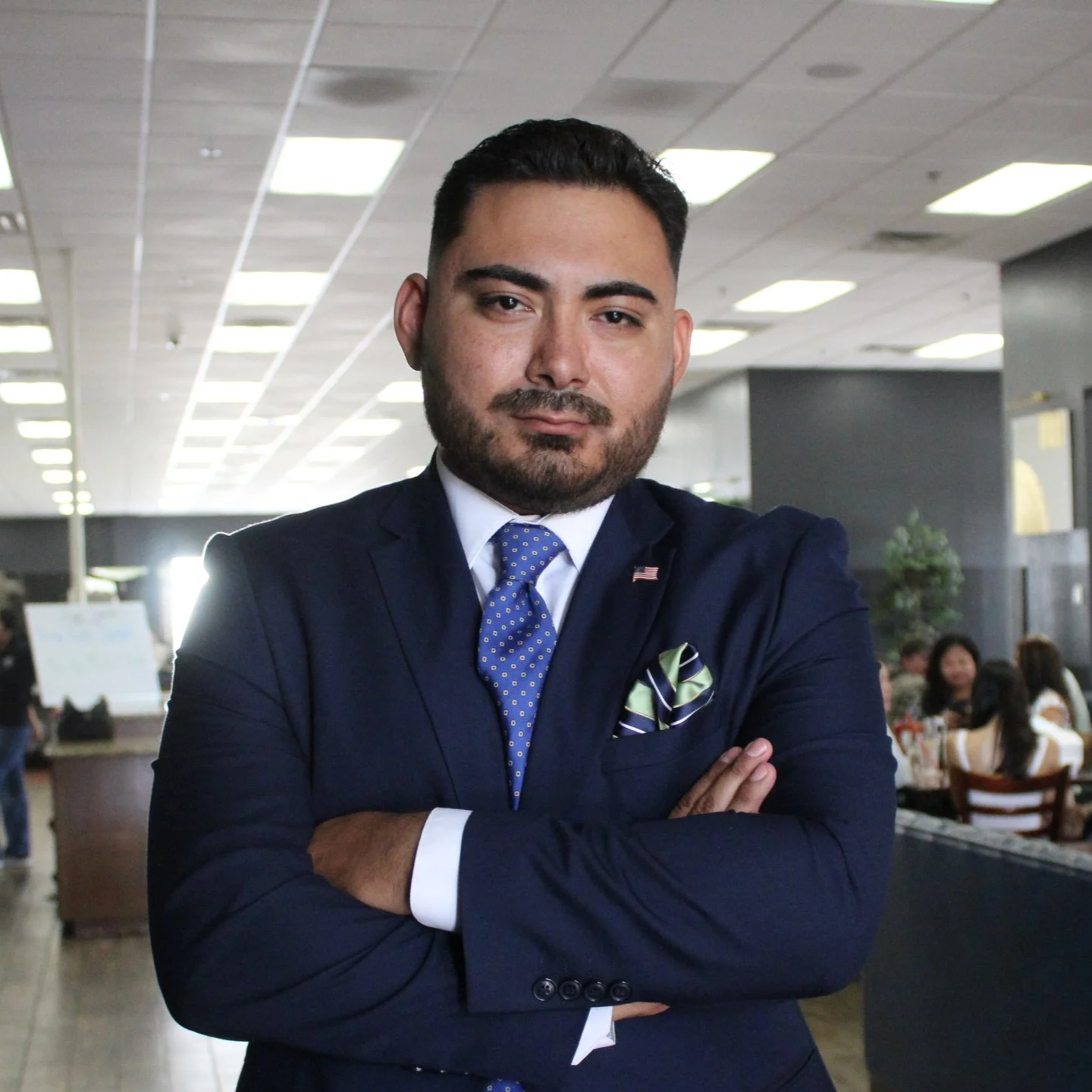 A confident man in a navy blue suit with a blue tie and a green patterned pocket square, standing with arms crossed inside a busy restaurant or cafe.