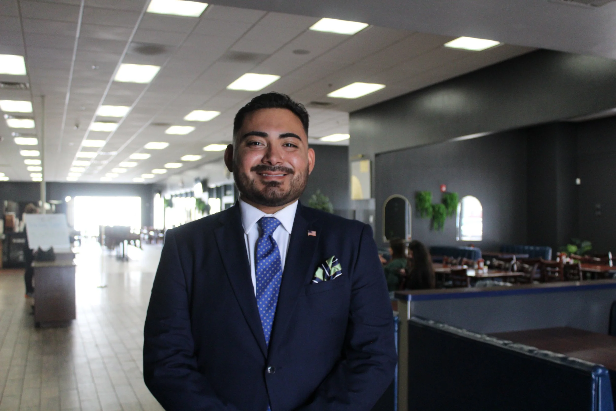 A man in a dark suit with a blue tie and pocket square smiling inside a well-lit restaurant or cafe.