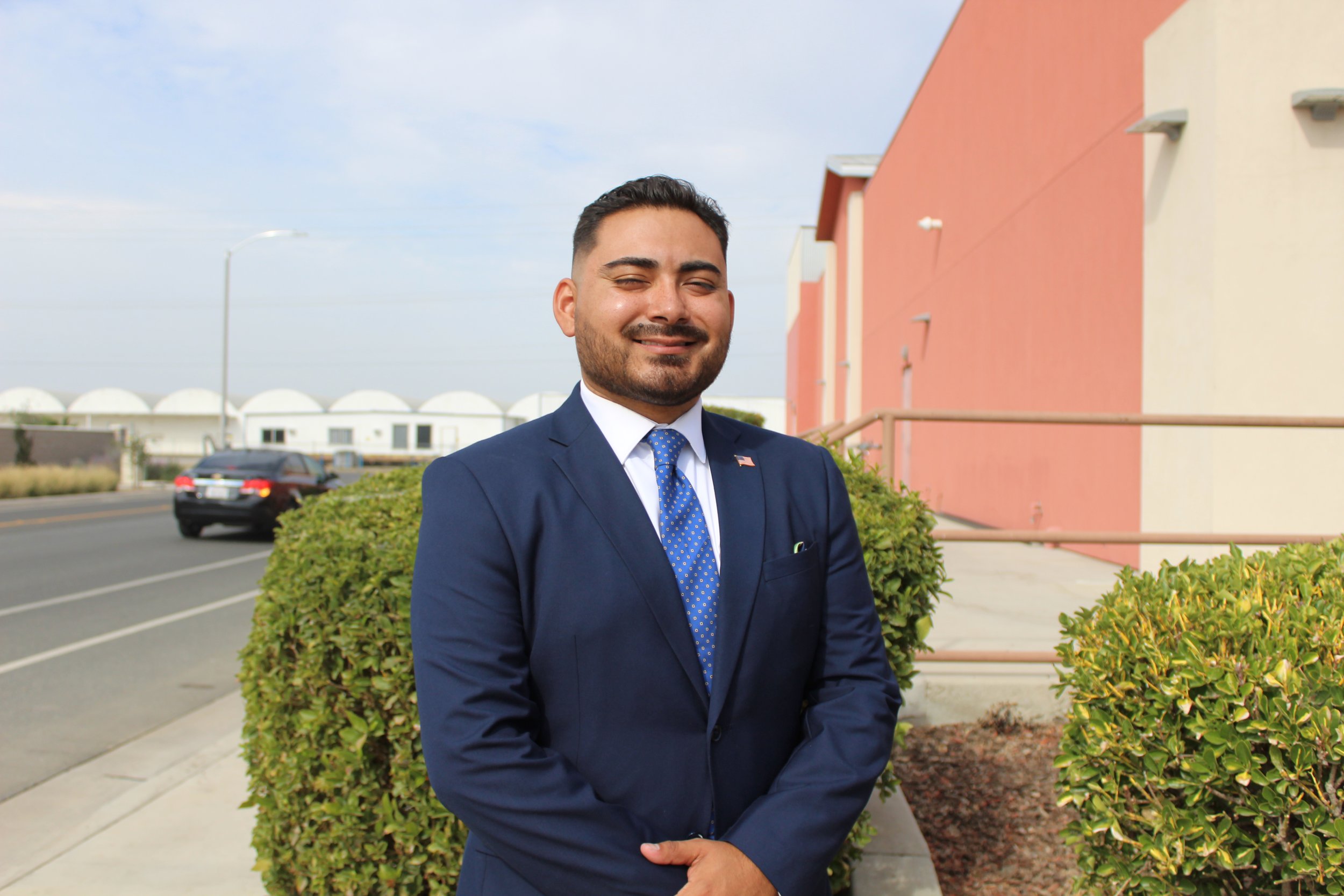 A man in a navy blue suit standing outdoors in front of a shrub, with a pink building and a parking lot in the background.
