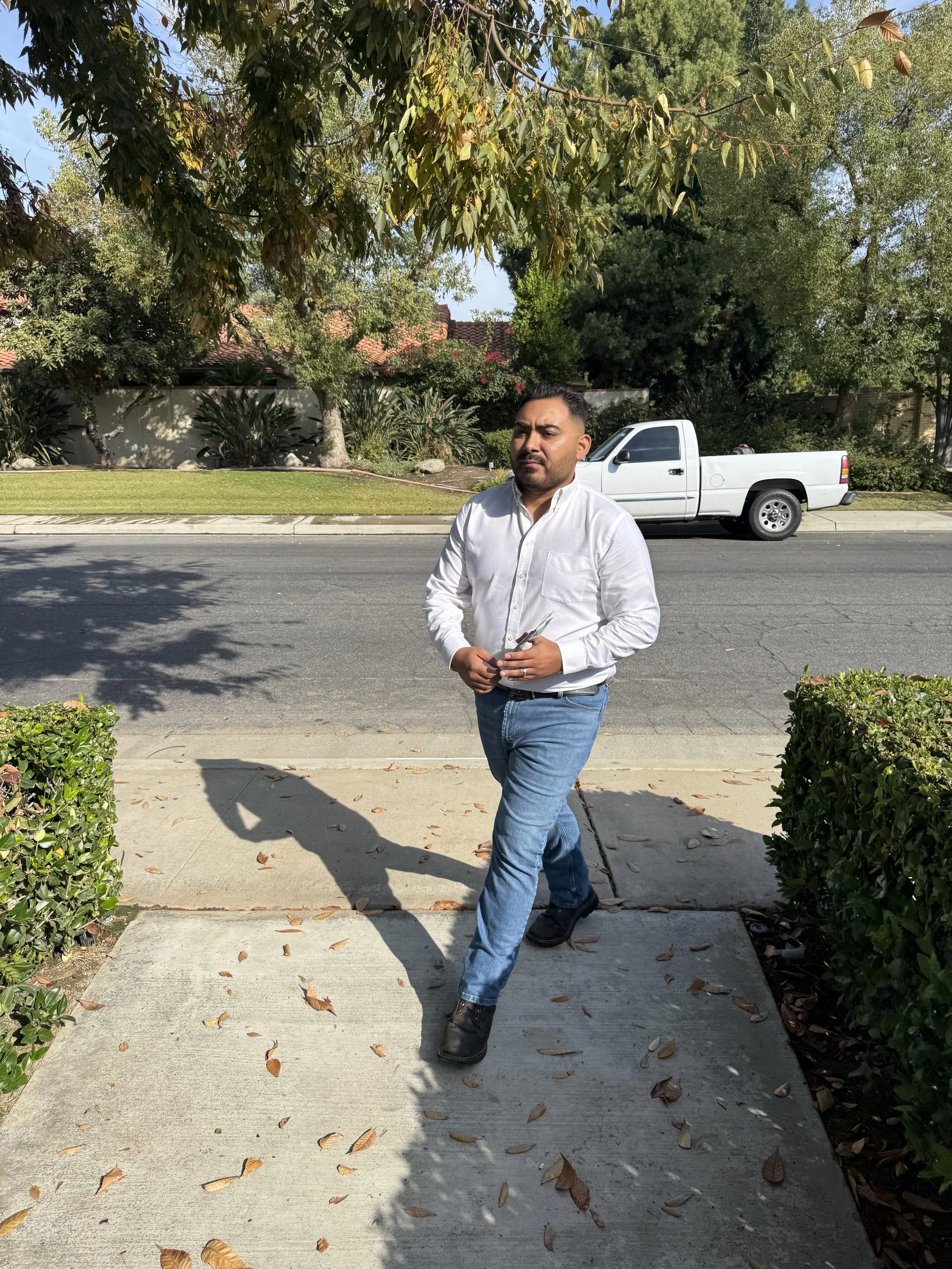 Man walking on sidewalk in front of a street, with trees and pickup truck in the background, and leaves on ground.