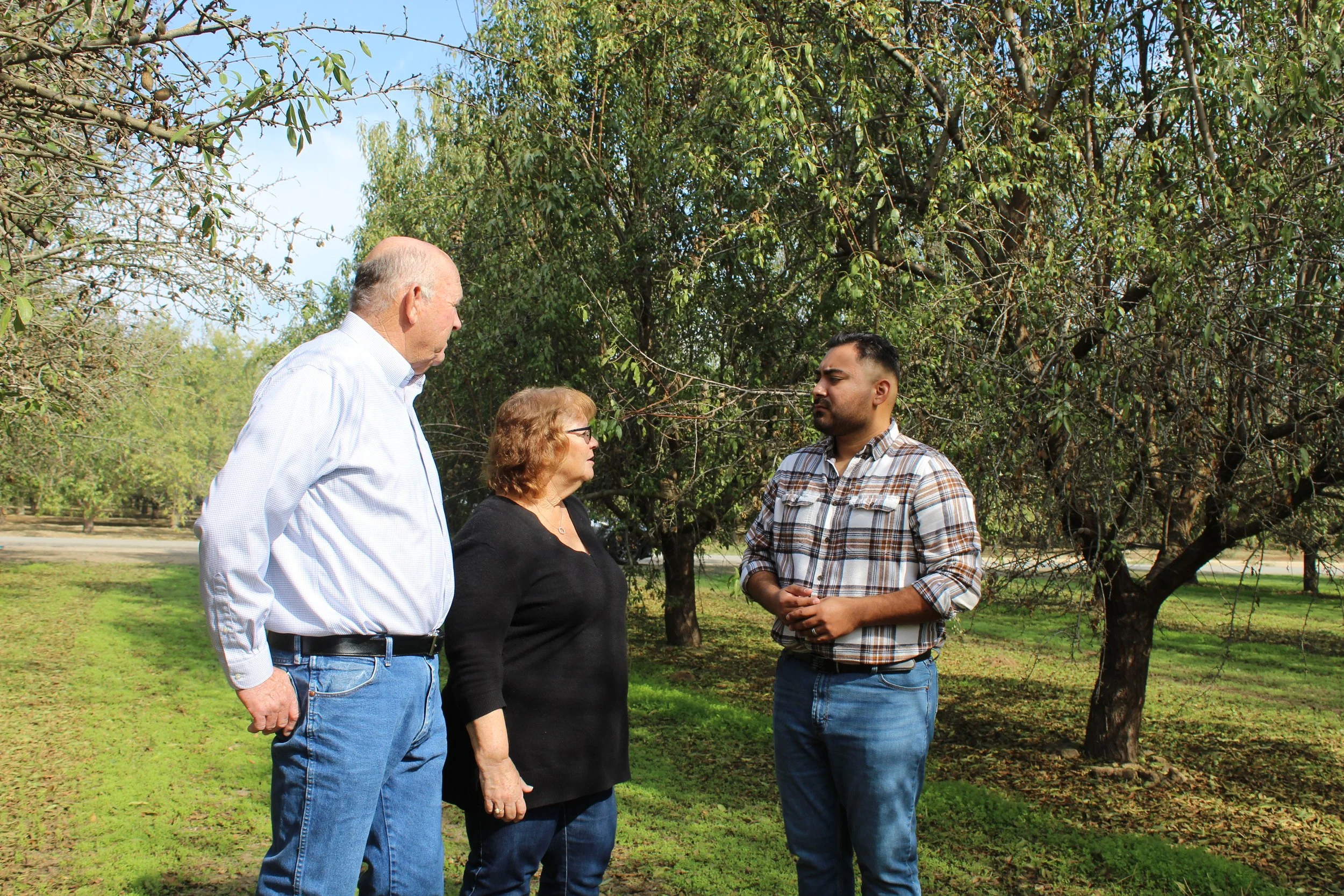 Three people standing outdoors under trees, engaged in a conversation. An older man in a light-colored shirt and jeans on the left, a middle-aged woman with glasses in the middle, and a younger man in a plaid shirt on the right.