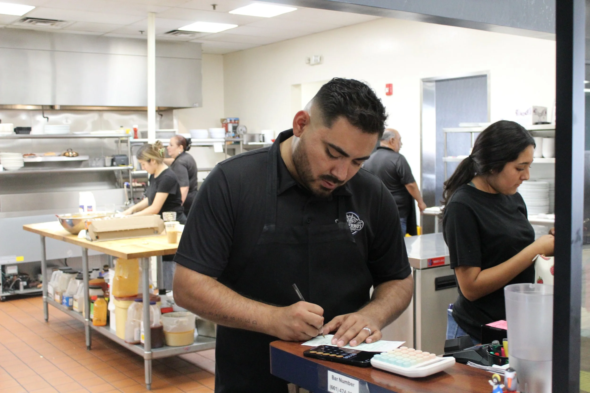 A man in a black shirt writes on a notepad while working behind a counter in a busy commercial kitchen. In the background, other staff members are preparing food in a well-organized kitchen with shelves and appliances.