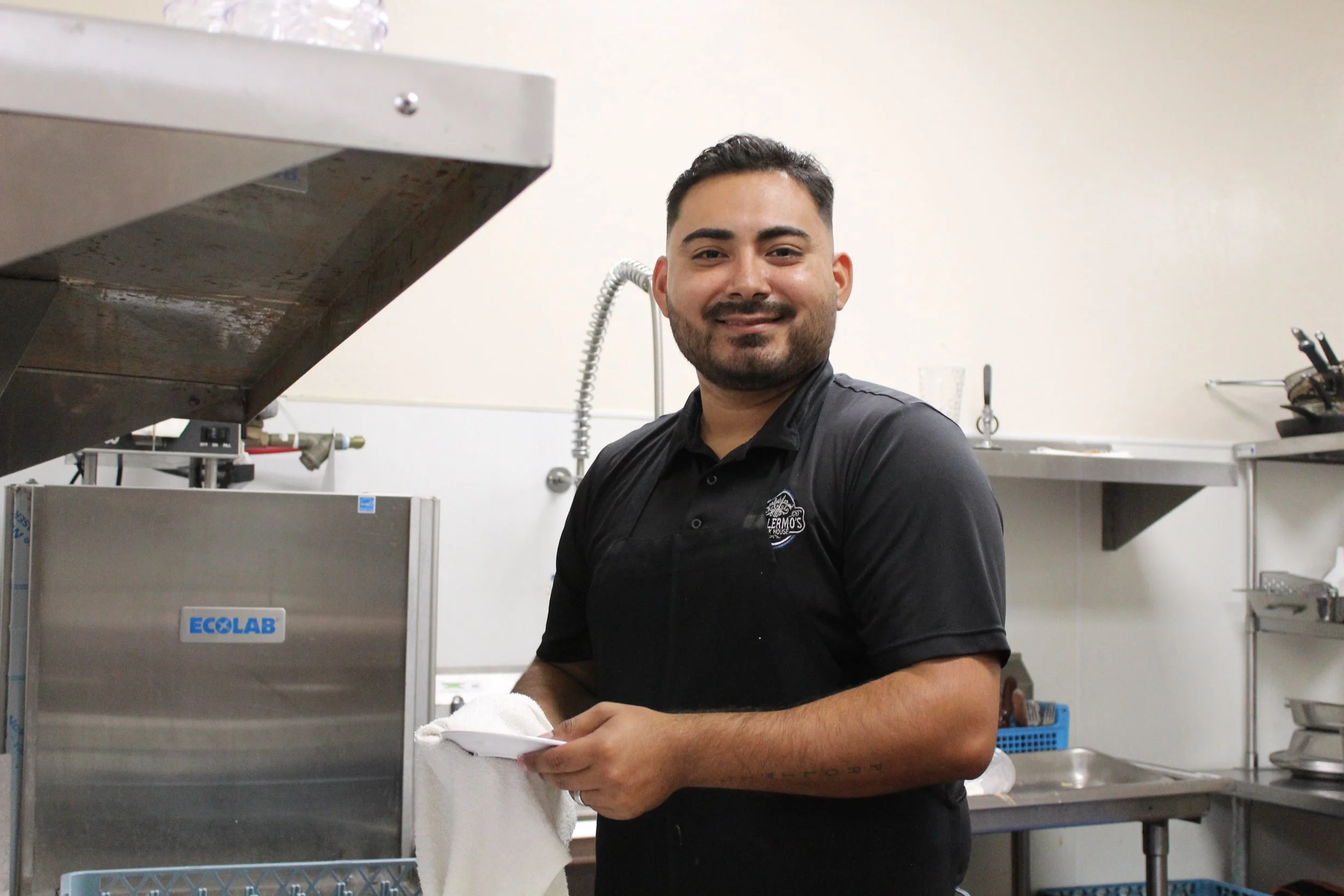 A smiling man with short dark hair and a beard, wearing a black shirt and apron, stands in a commercial kitchen holding a towel and a small plate.