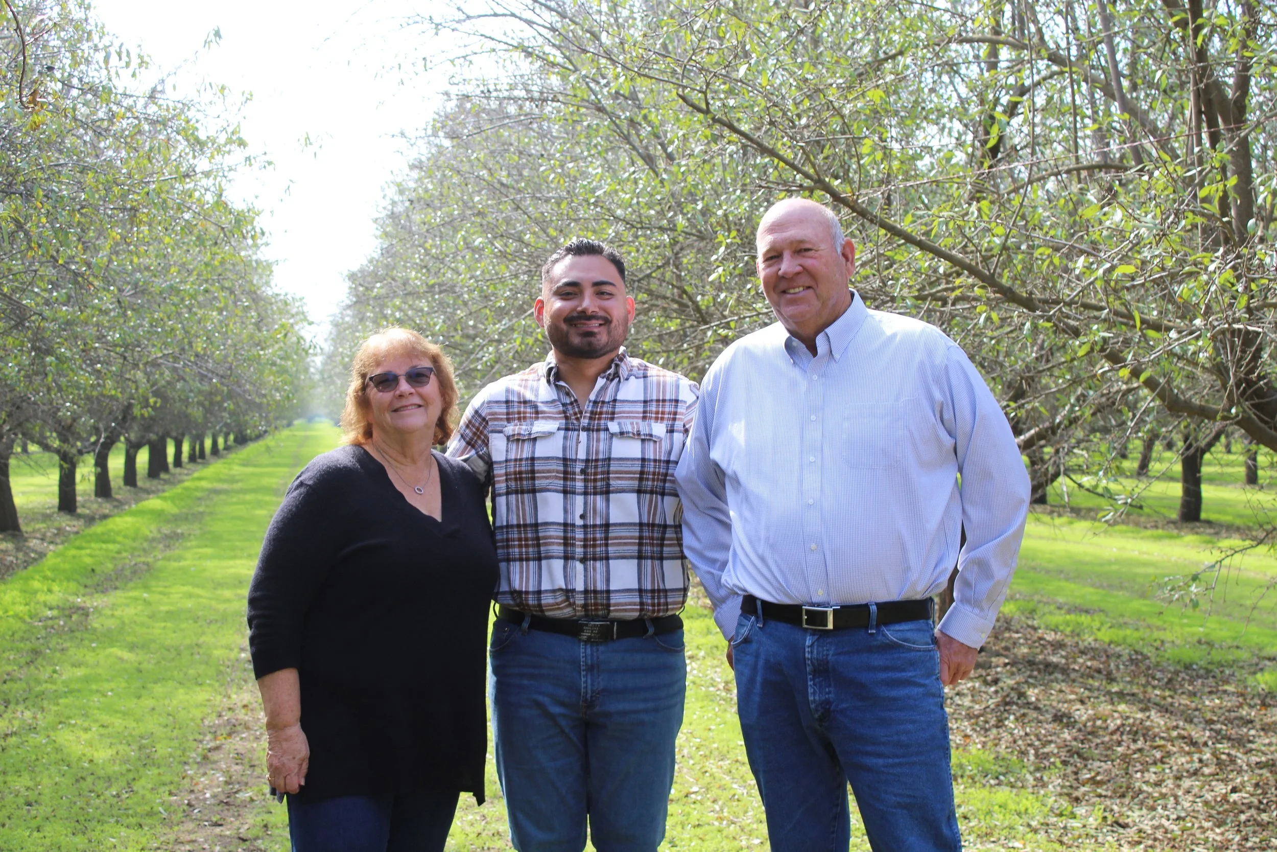 Three people, two men and one woman, standing in an orchard with blooming trees, smiling at the camera during daytime.