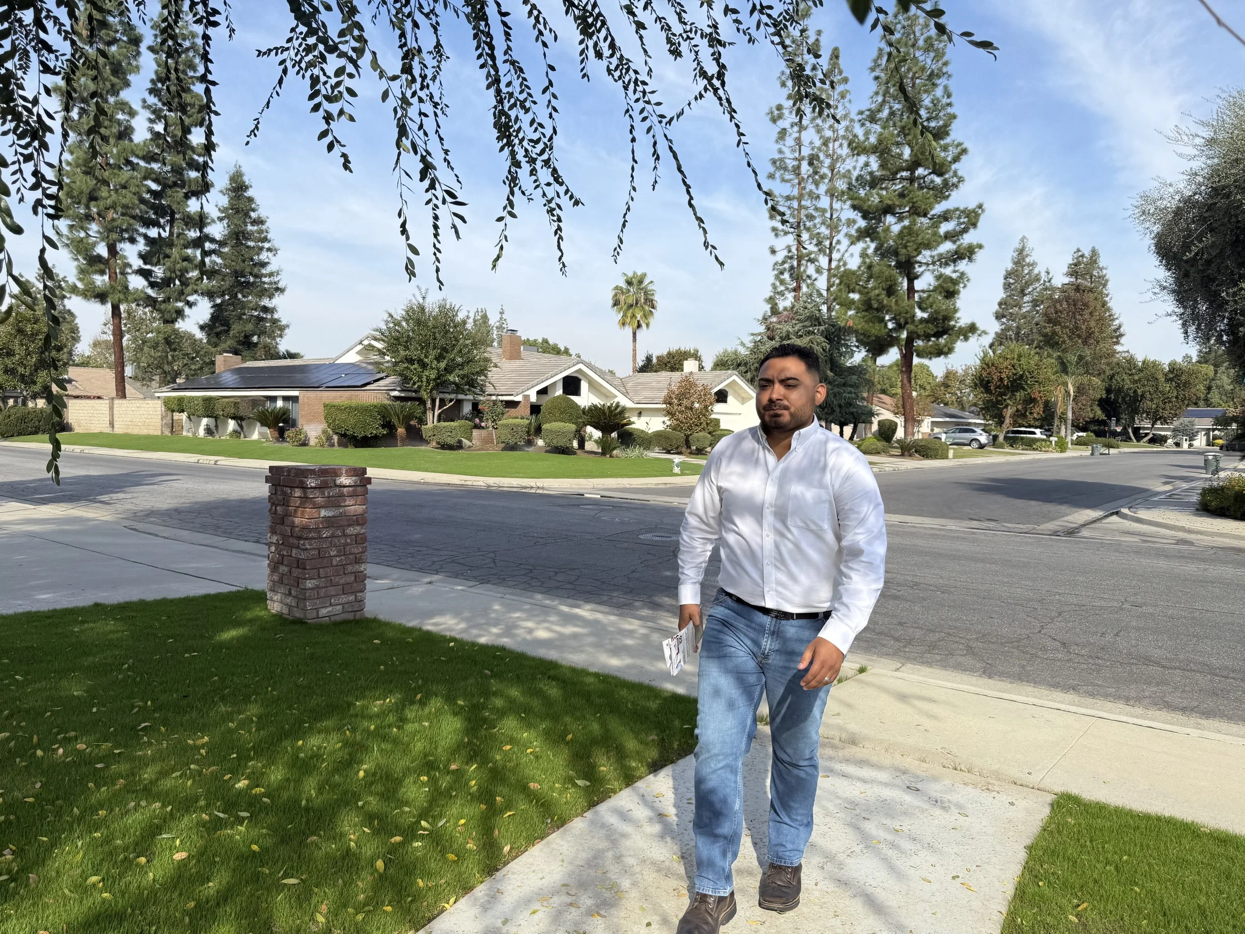 A man walking on a sidewalk in a suburban neighborhood with green lawns and houses, some with solar panels, trees, and a blue sky.