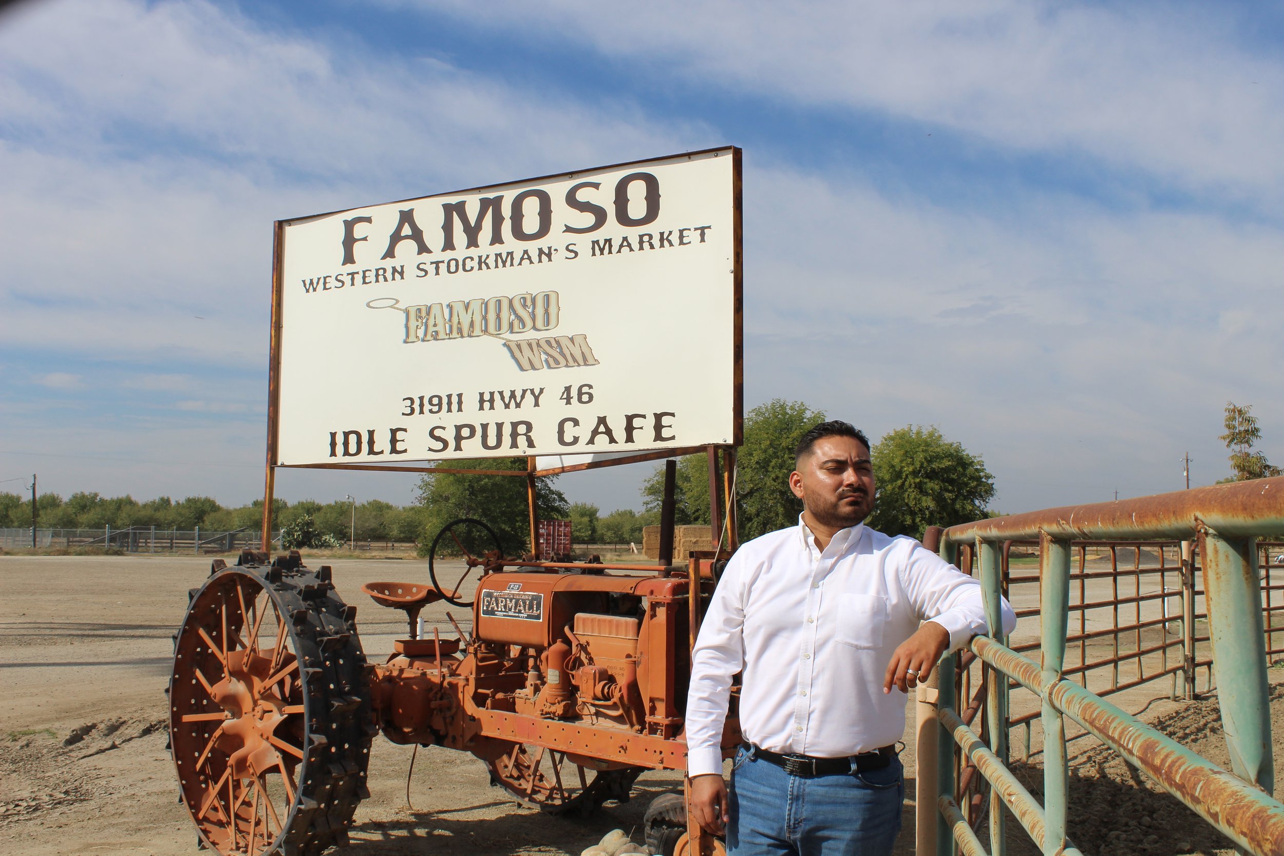 A man in a white shirt leaning on a metal fence in front of an old tractor with a large sign overhead advertising FAMSO Western Stockman's Market, located at 31911 Hwy 46, Idle Spur Cafe, with a background of an open, rural landscape under a partly c