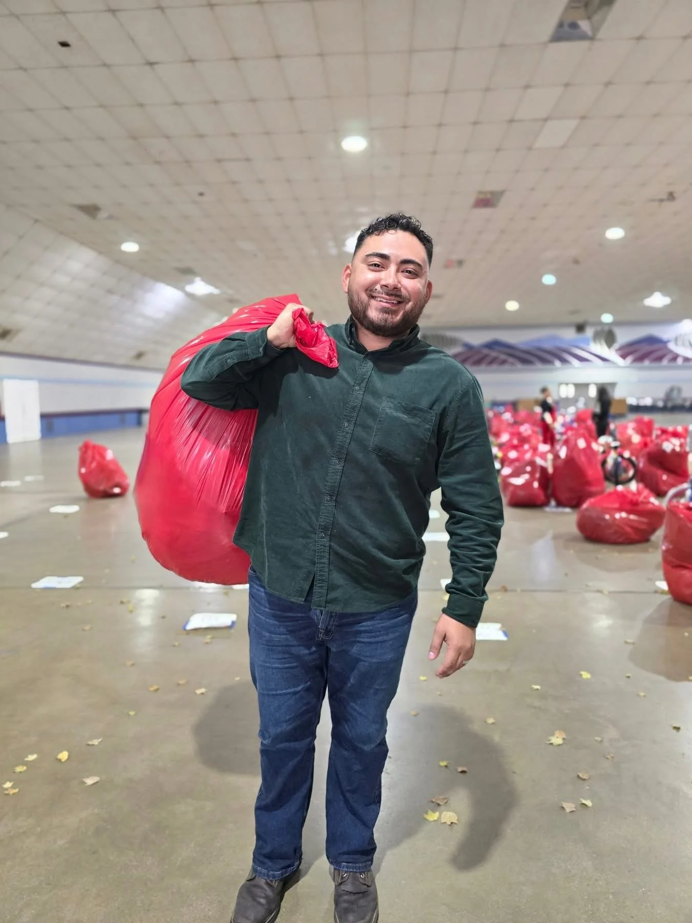 A man holding a large red plastic bag over his shoulder, standing in an indoor space with other red bags in the background.