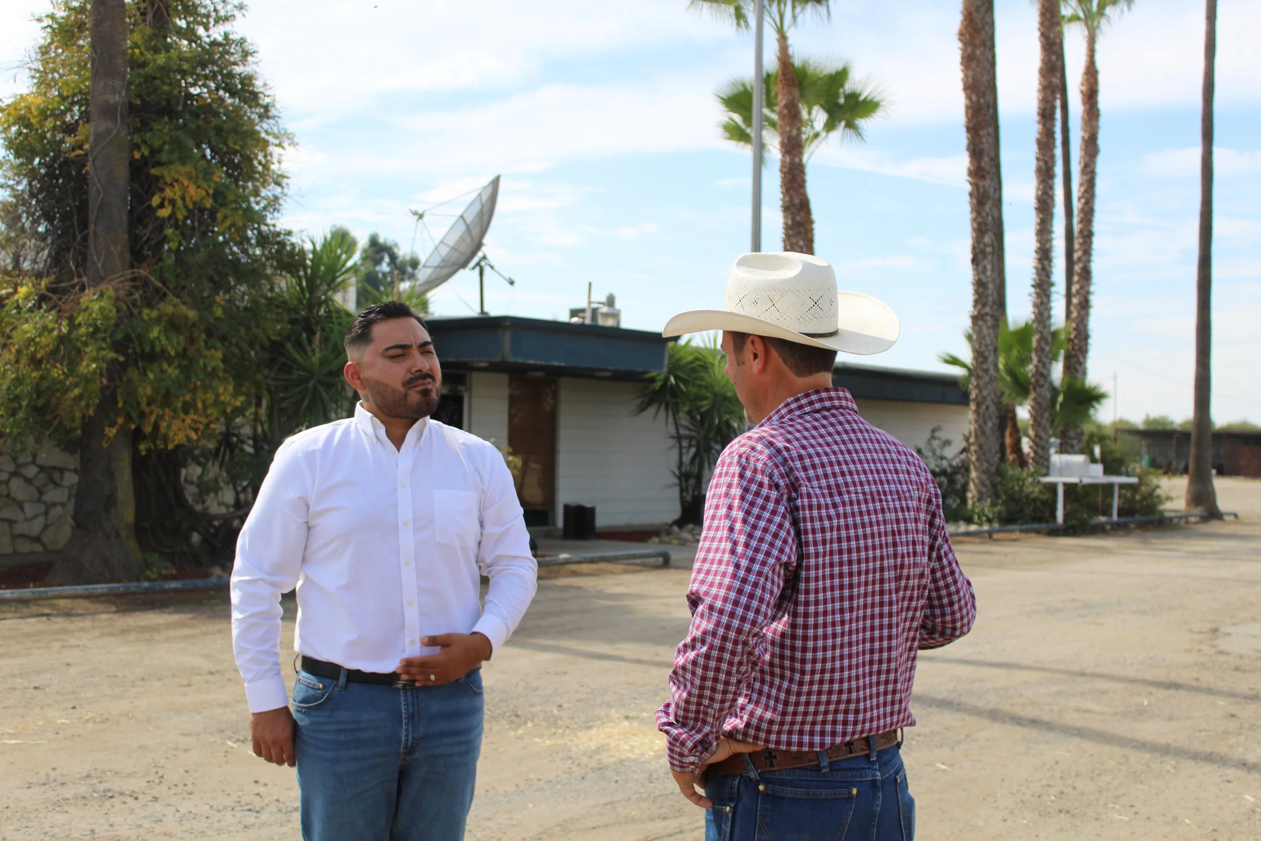 Two men standing and talking outdoors, one wearing a white shirt and jeans, the other wearing a plaid shirt, jeans, and a white cowboy hat, with palm trees and a building in the background.