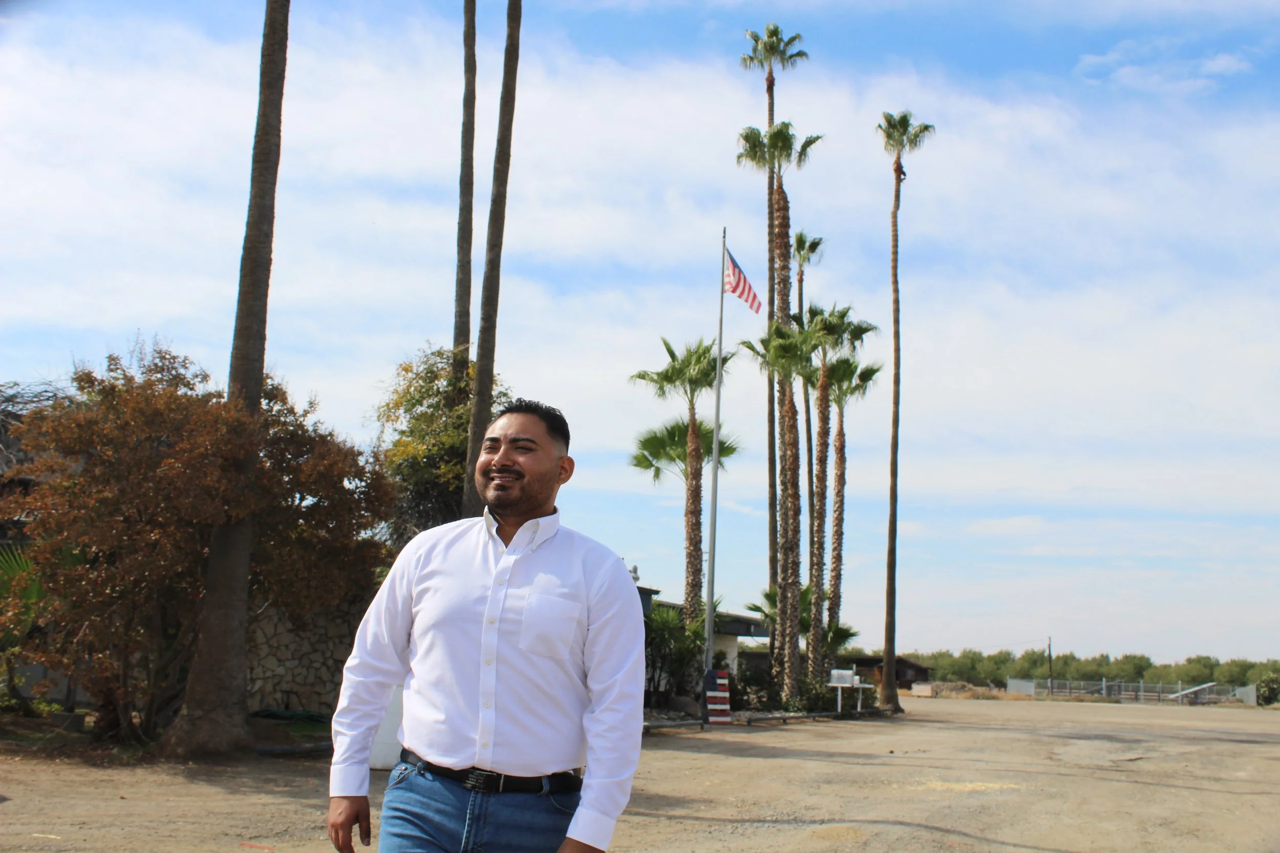 A man in a white shirt and blue jeans standing outdoors with palm trees and an American flag in the background on a partly cloudy day.