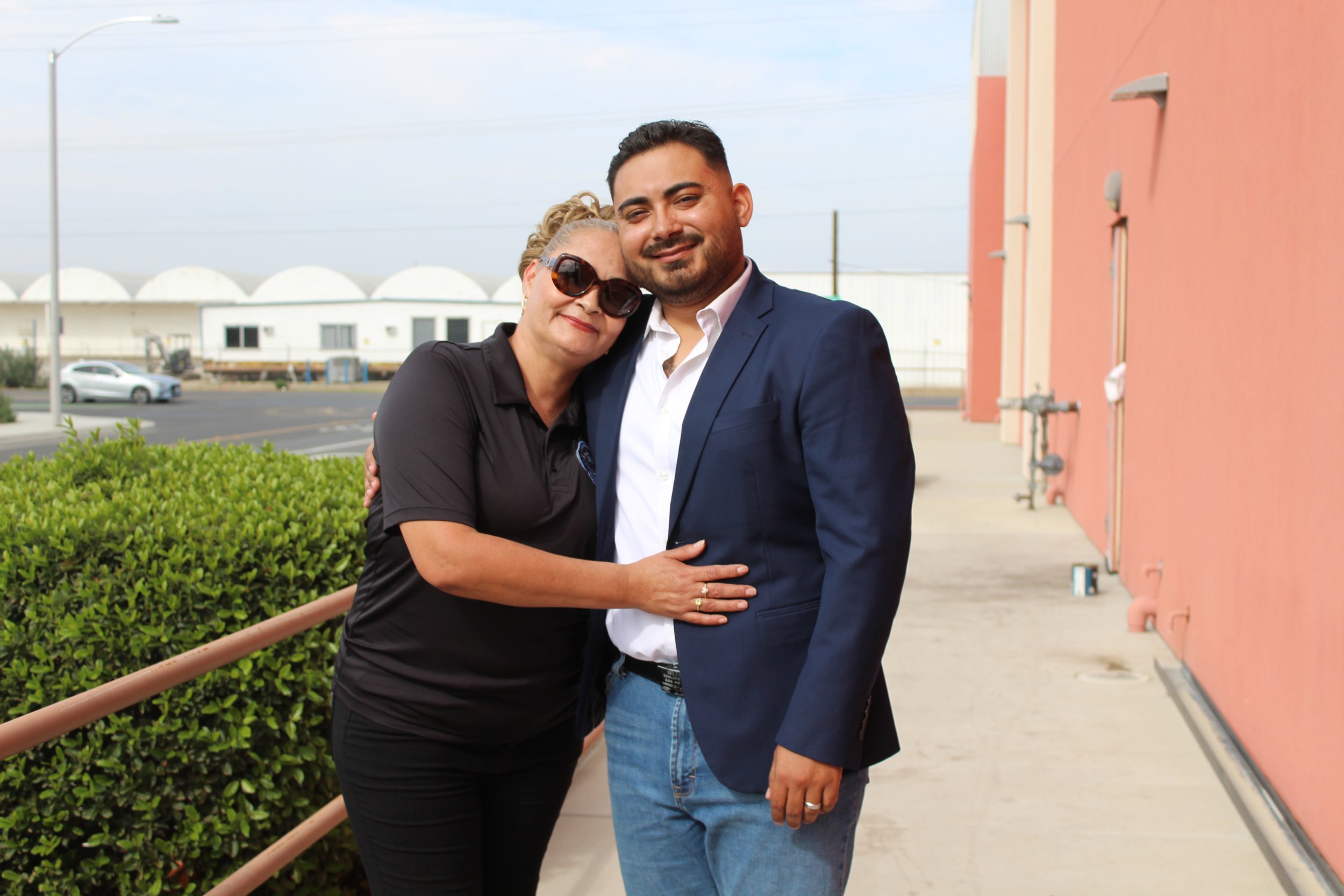 A woman in black polo shirt and sunglasses hugging a man in a blue blazer and jeans outside near a pink building, with a parking lot in the background.