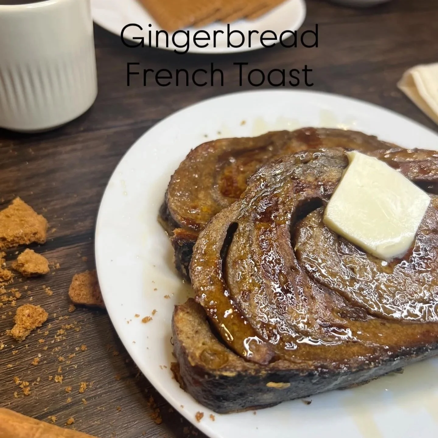 Close-up of a caramelized cinnamon roll with a pat of butter on top, served on a white plate. The background includes a jar and some crumbs on a wooden table, with the text 'Gingerbread French Toast' overlaid.