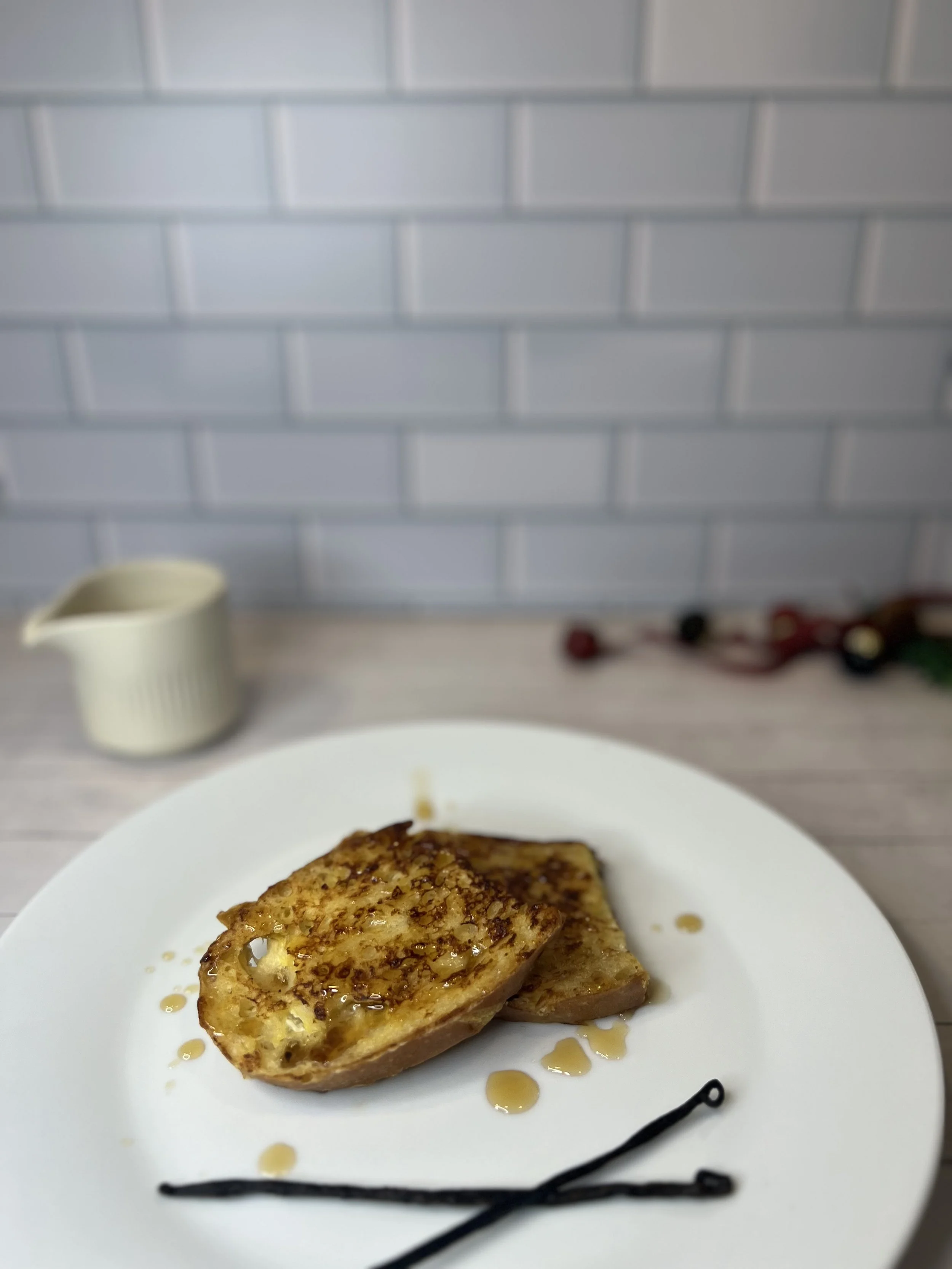 Two slices of golden-brown vanilla bean French toast made with sourdough, drizzled with maple syrup, served on a white plate with whole vanilla beans in the foreground. A small cream pitcher and holiday décor sit softly blurred in the background.