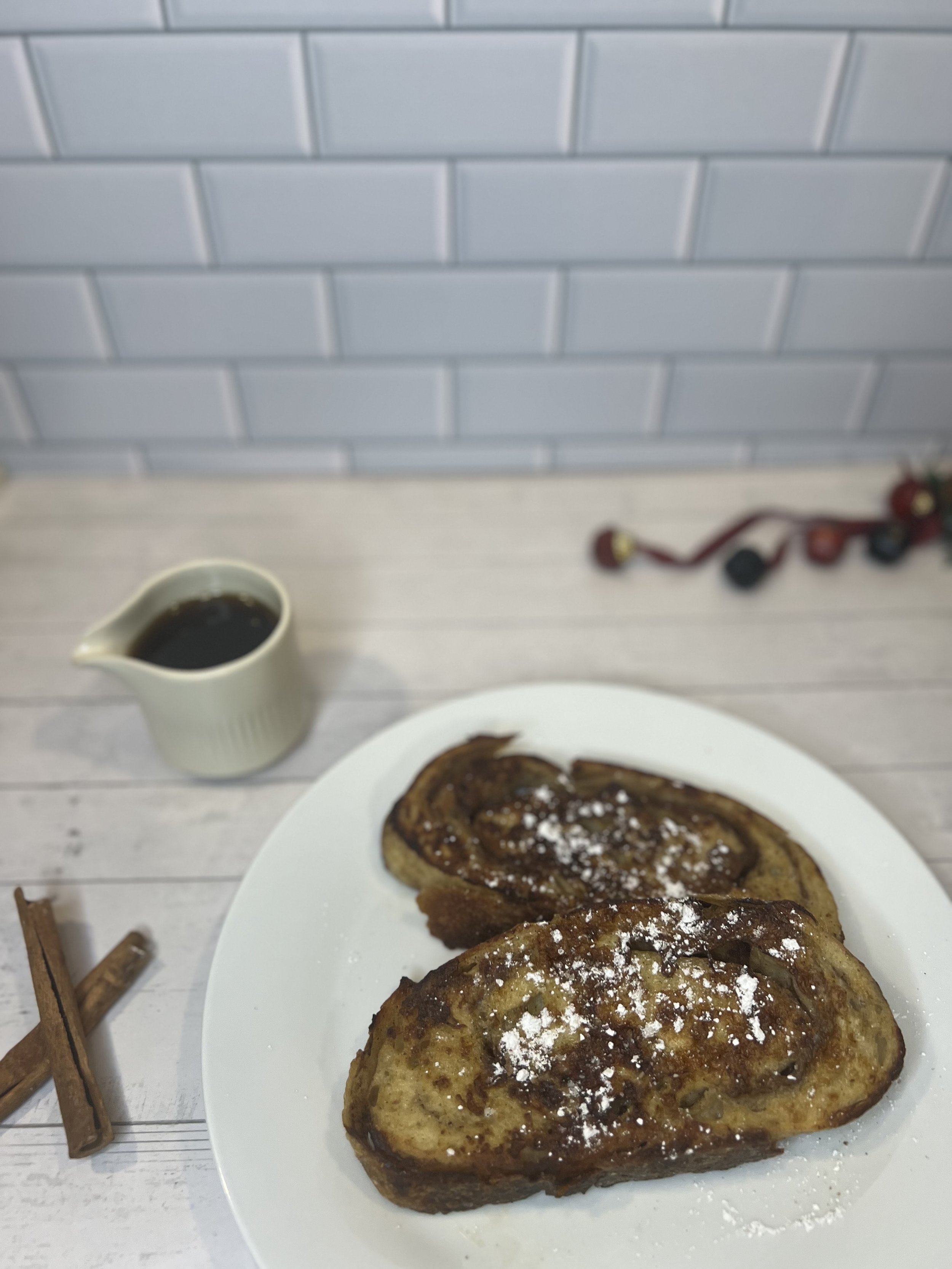 A white plate with two cinnamon rolls topped with powdered sugar, accompanied by a small pitcher of syrup, on a light-colored wooden table with cinnamon sticks nearby, against a background of white subway tiles.