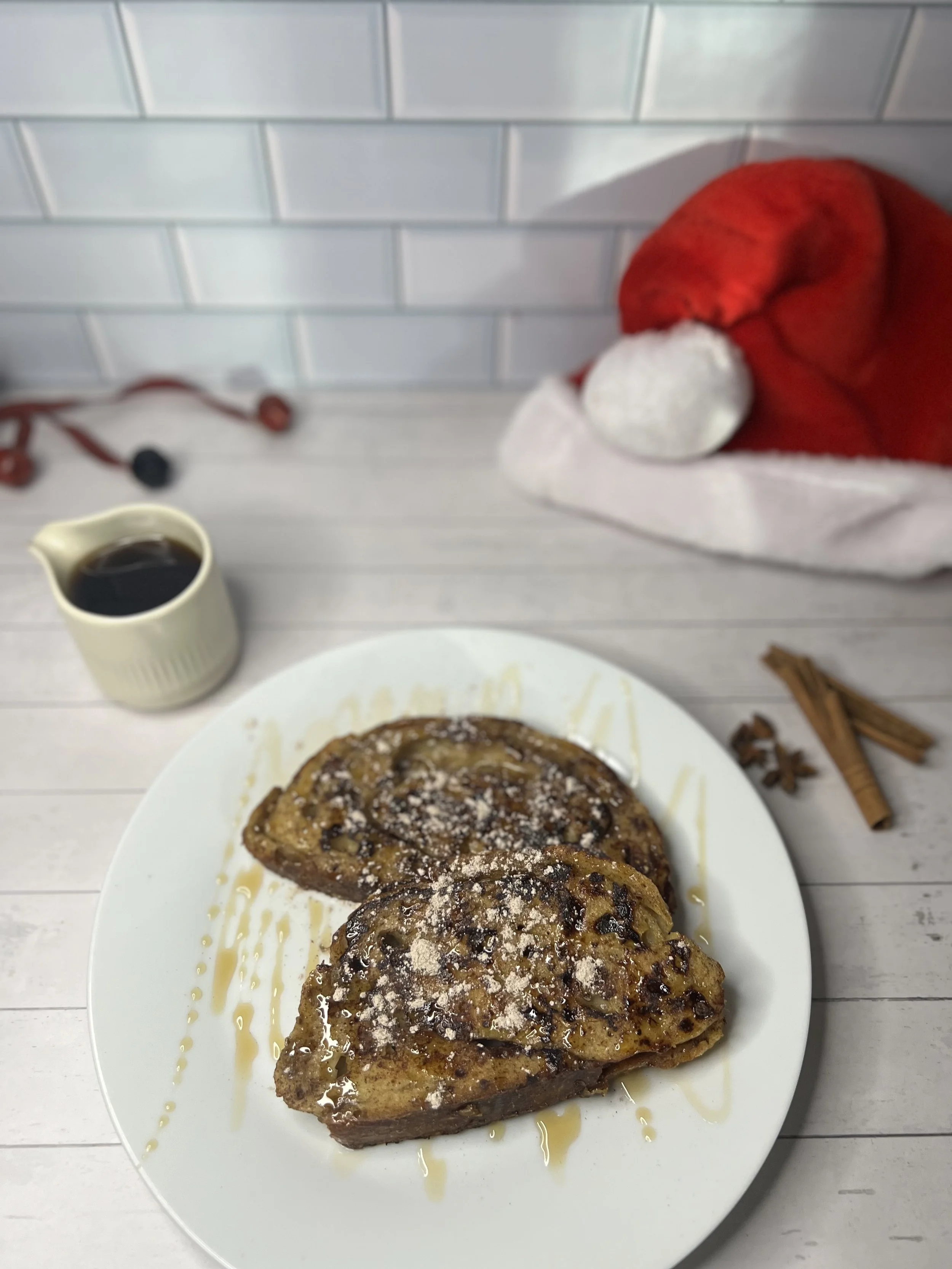 A white plate with two heart-shaped French toasts drizzled with syrup and sprinkled with powdered sugar, a small cream pitcher with syrup, a Santa hat, cinnamon sticks, and a pair of glasses on a white wooden table against a white tile backsplash.