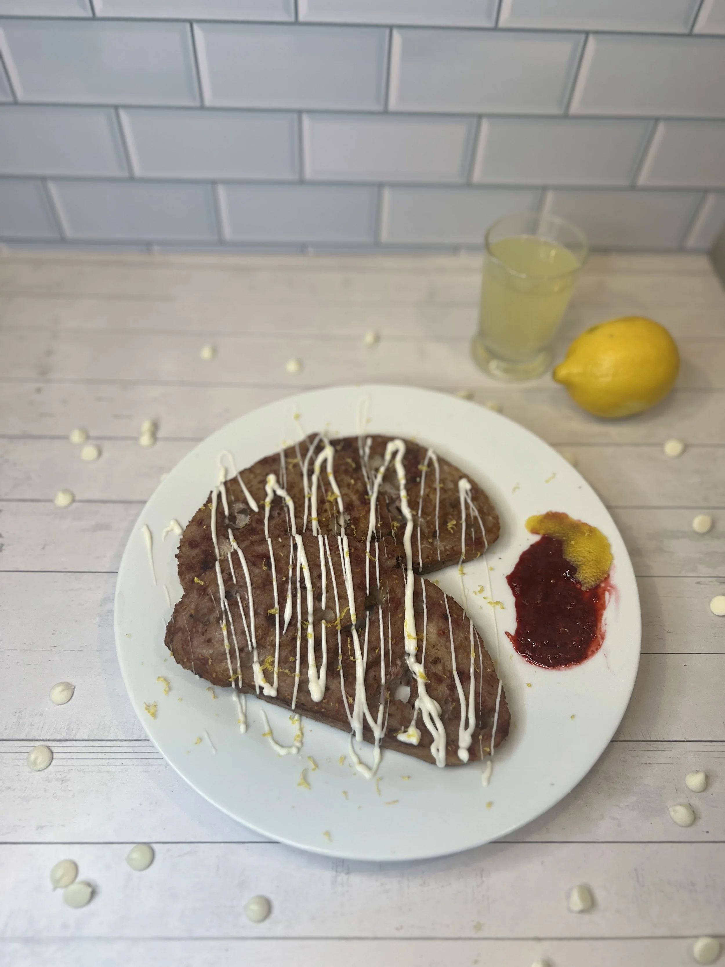 Stack of raspberry lemon French toast made with sourdough, drizzled with white chocolate and lemon zest, served with a raspberry sauce streak. A lemon and a glass of lemon drink sit blurred in the background.