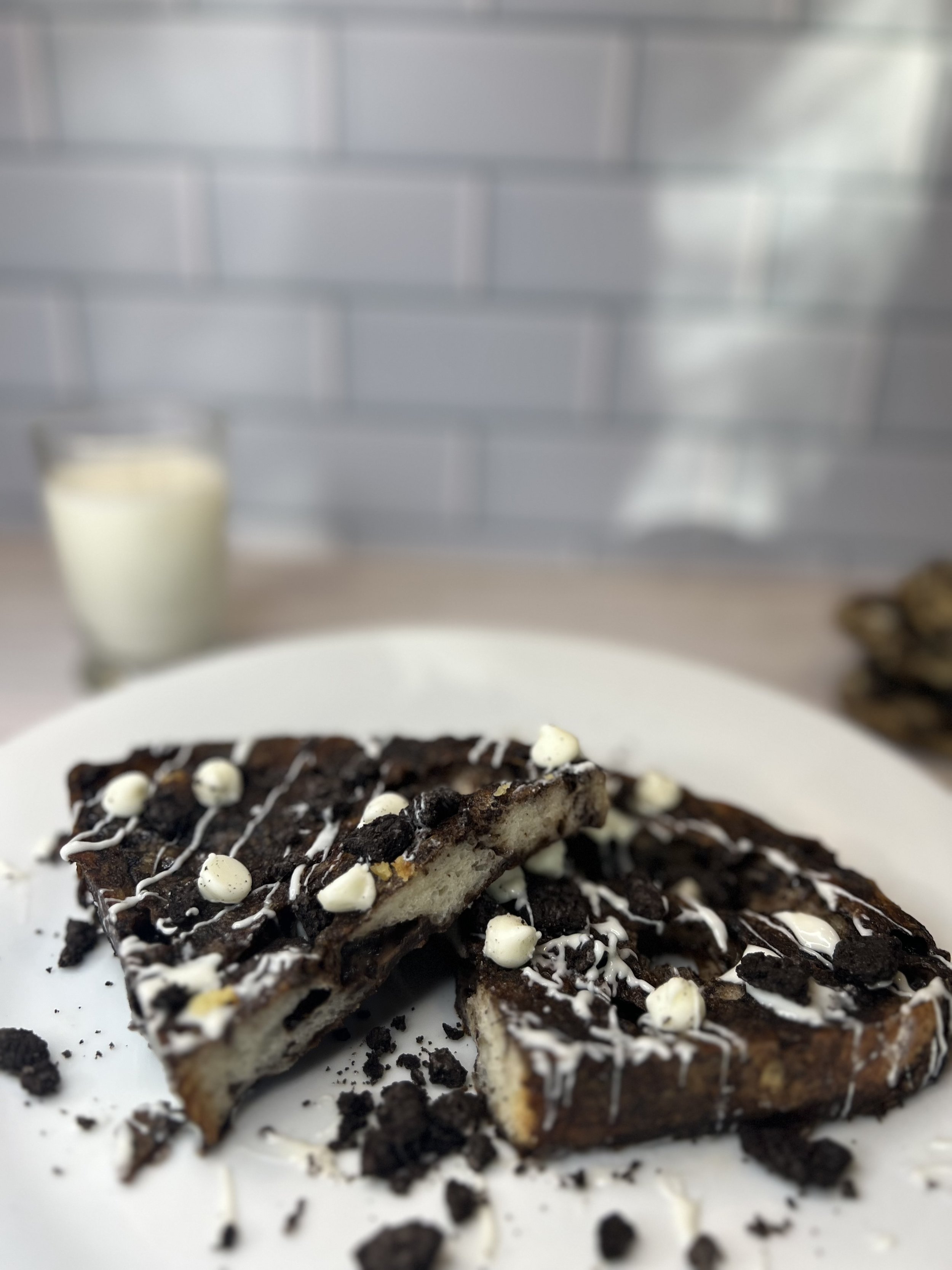 Cookies & Cream French toast on a white plate, topped with crushed chocolate cookies, white chocolate drizzle, and white chocolate chips. A glass of milk sits blurred in the background on a light wood surface with a subway tile backdrop.