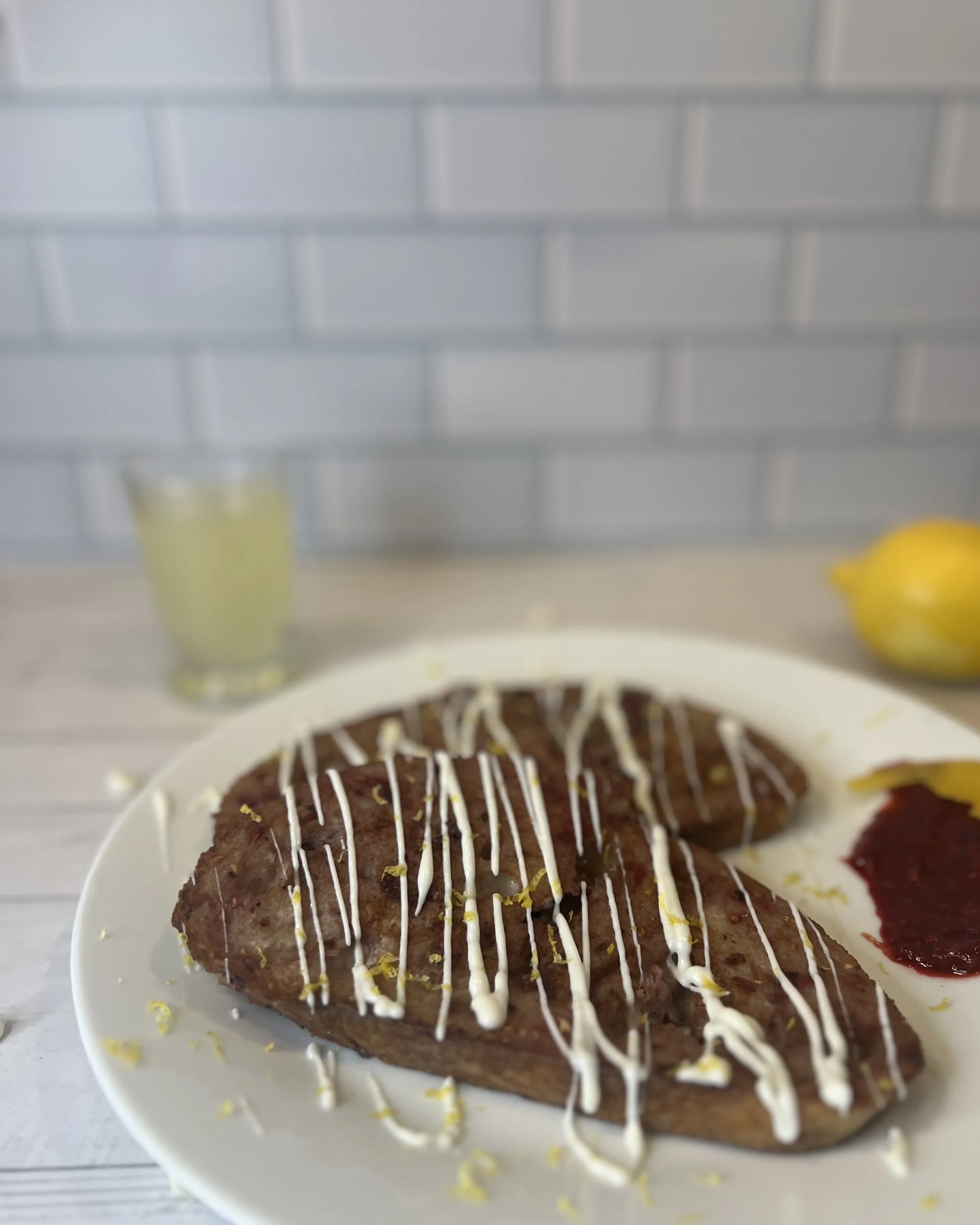 A plate with grilled steak, topped with white sauce drizzle, accompanied by lemon wedges and a side of red sauce. A lemon and a glass of lemonade are in the background on a wooden table, with a brick wall behind.