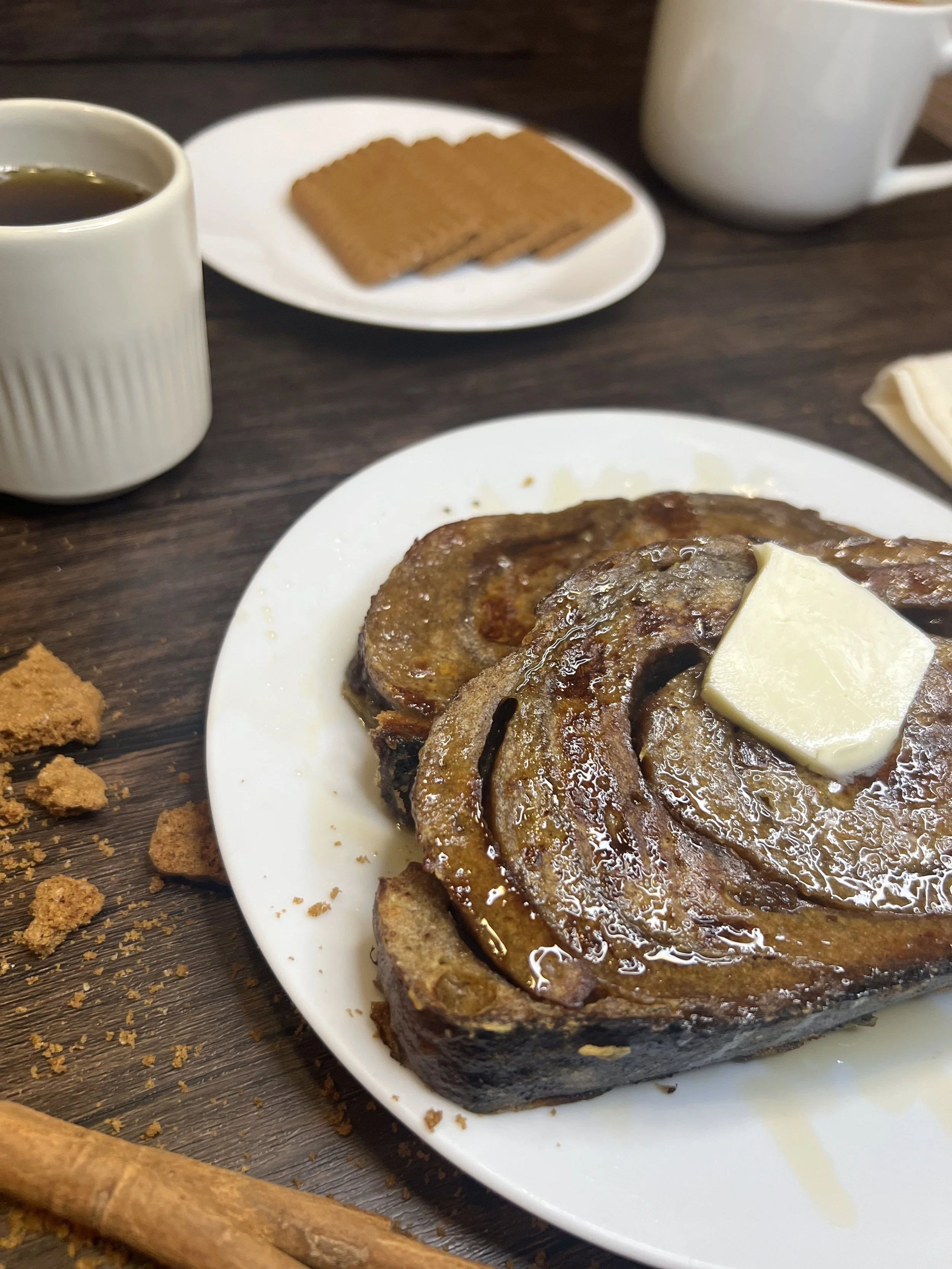 Close-up of thick-cut gingerbread sourdough French toast topped with a melting pat of butter and drizzled lightly with syrup, showing the warm cinnamon swirls and caramelized edges on a white plate.
