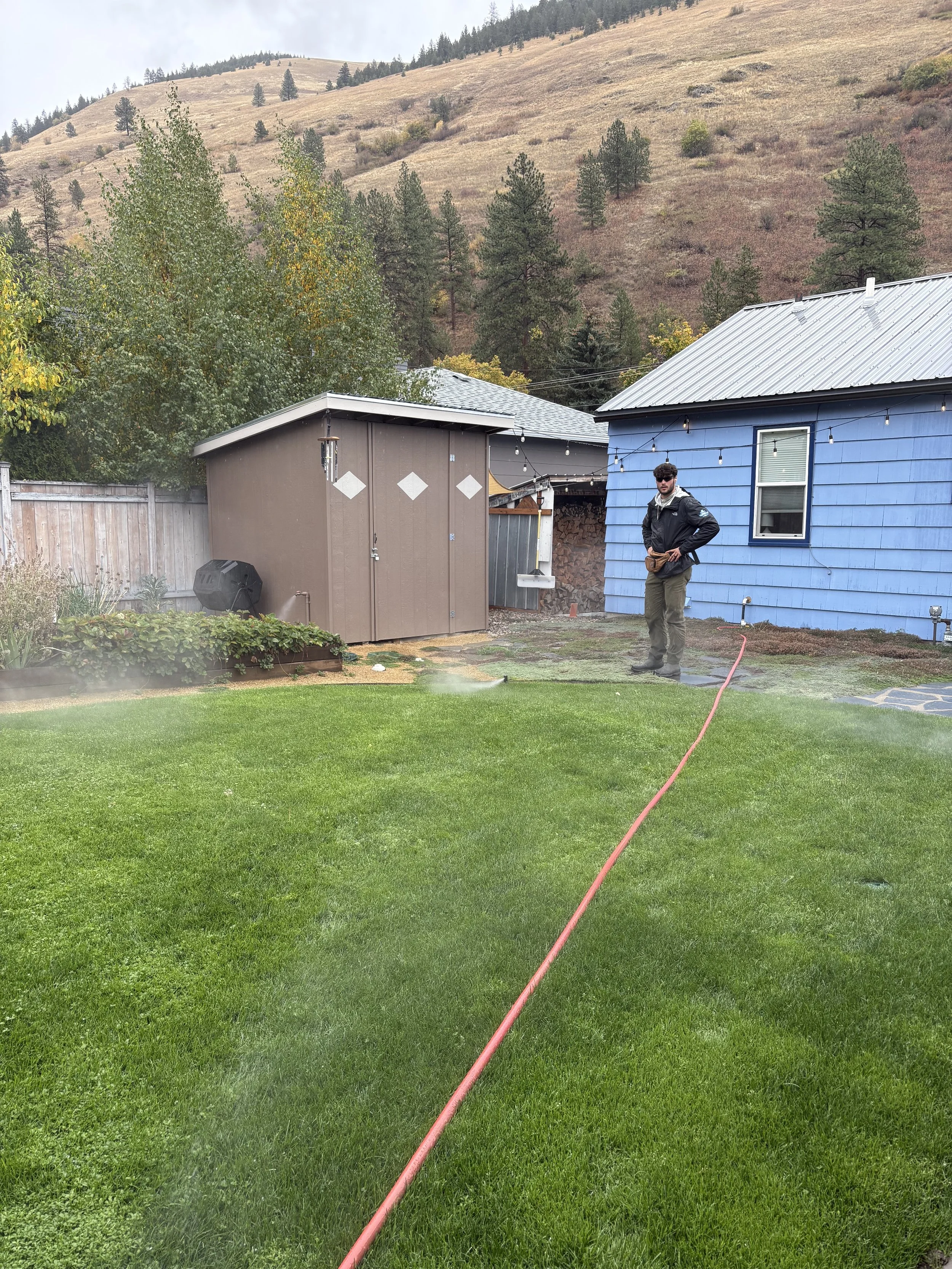 A man standing on a lawn next to an air compressor's hose, blowing out the sprinkler system in order that the sprinkler system will be properly winterized.