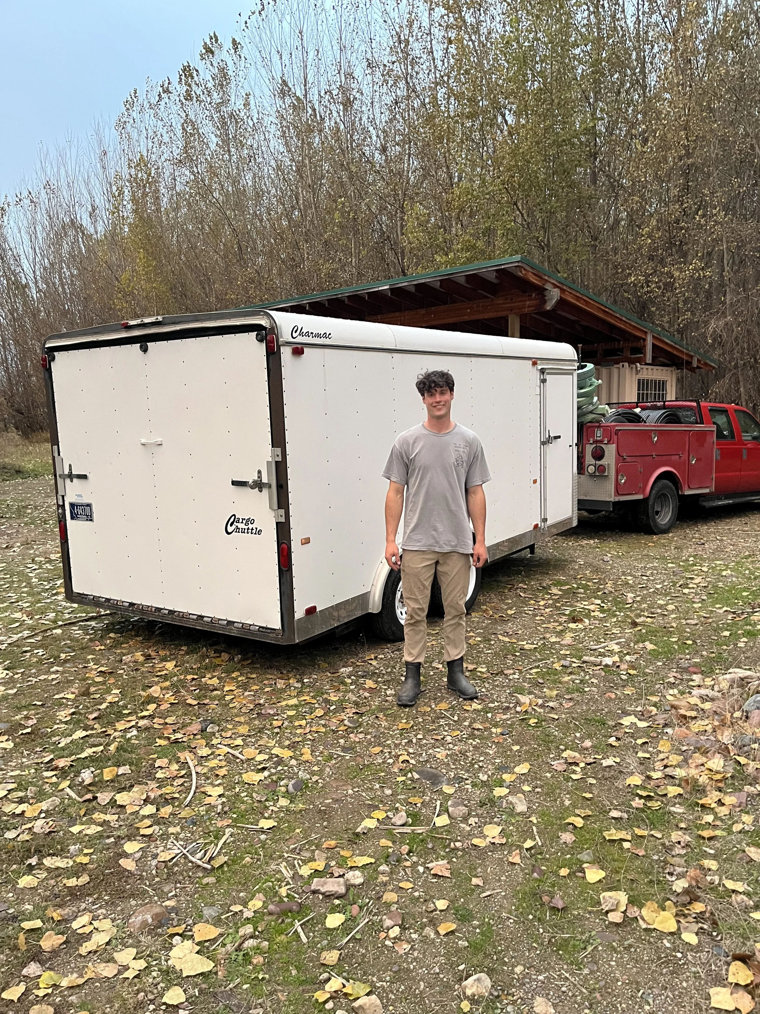 A young man stands in front of a white cargo trailer attached to a red truck, which are used for his sprinkler company.