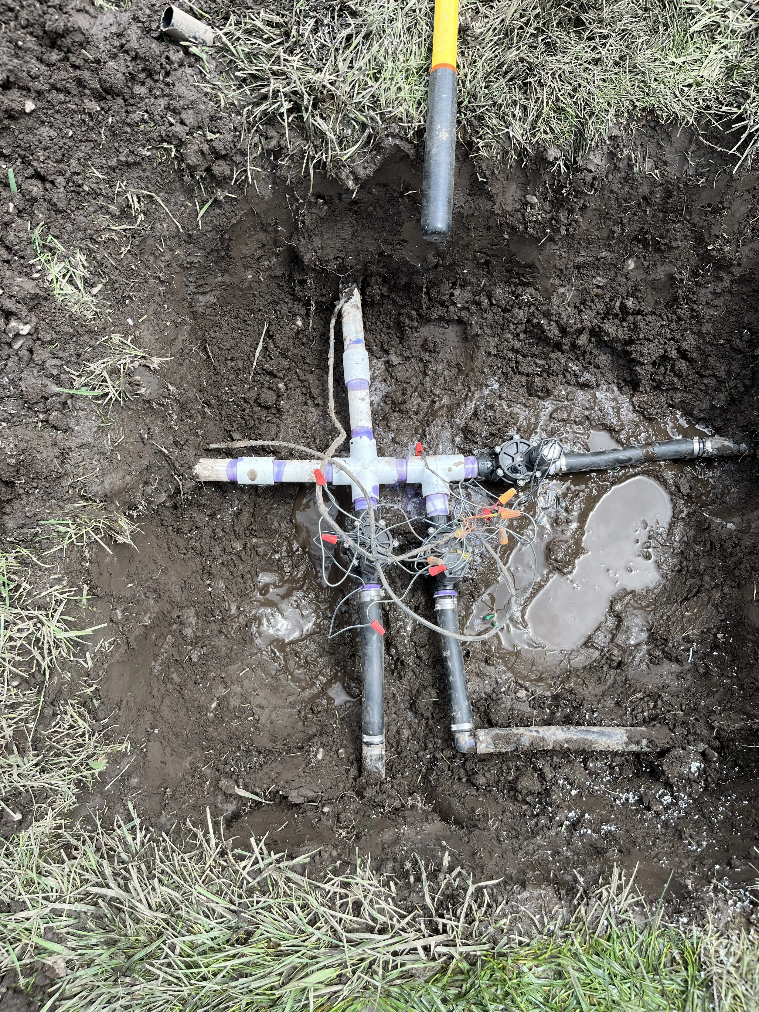 Underground utility pipes and wiring with exposed fittings and valves in a muddy excavation site surrounded by grass, including a yellow-handled tool at the top.