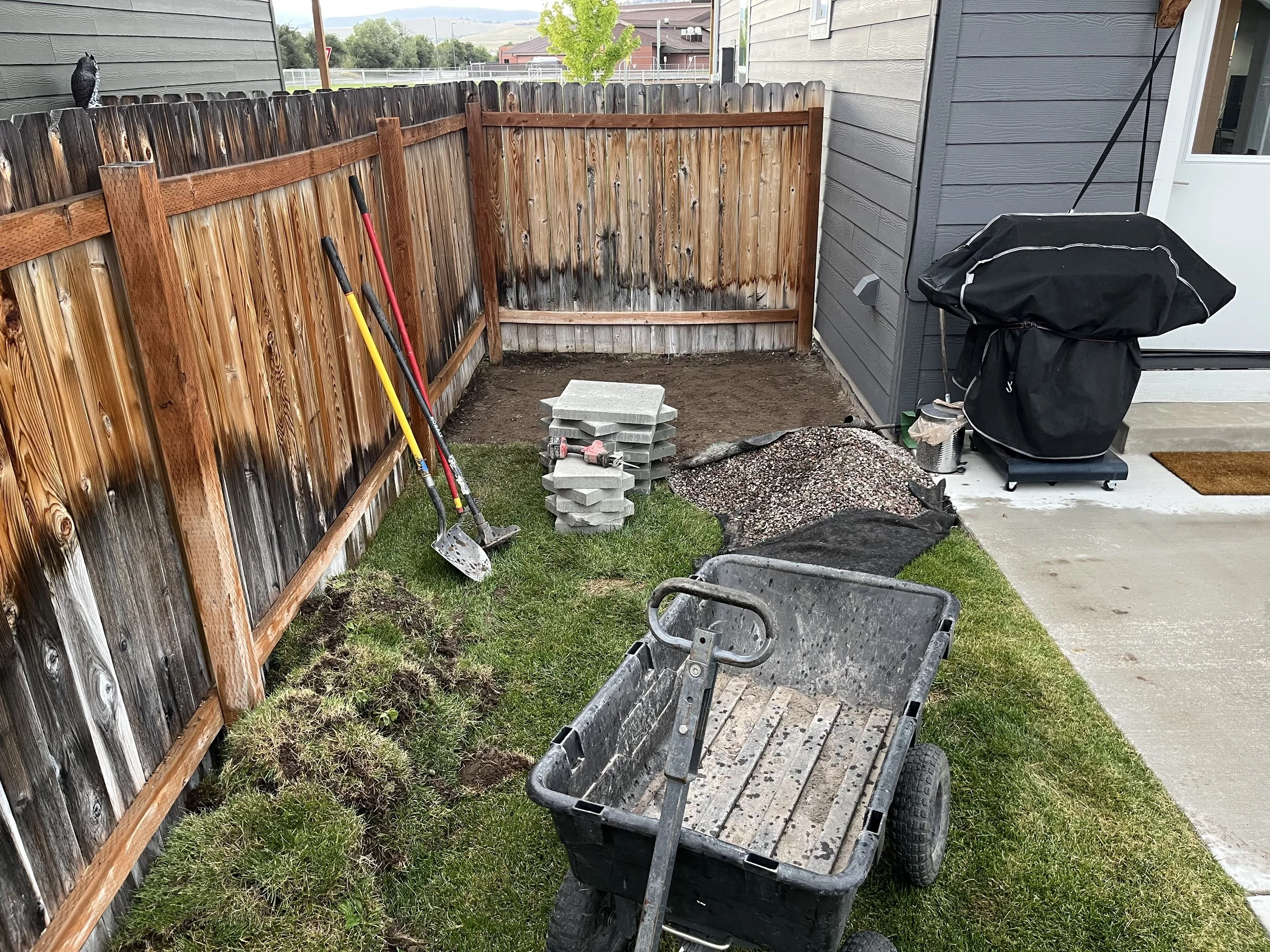 Backyard with a wooden fence, gardening tools, stack of concrete pavers, piles of gravel and soil, a wheelbarrow, and a covered grill near a house, showcasing a sprinkler installation.