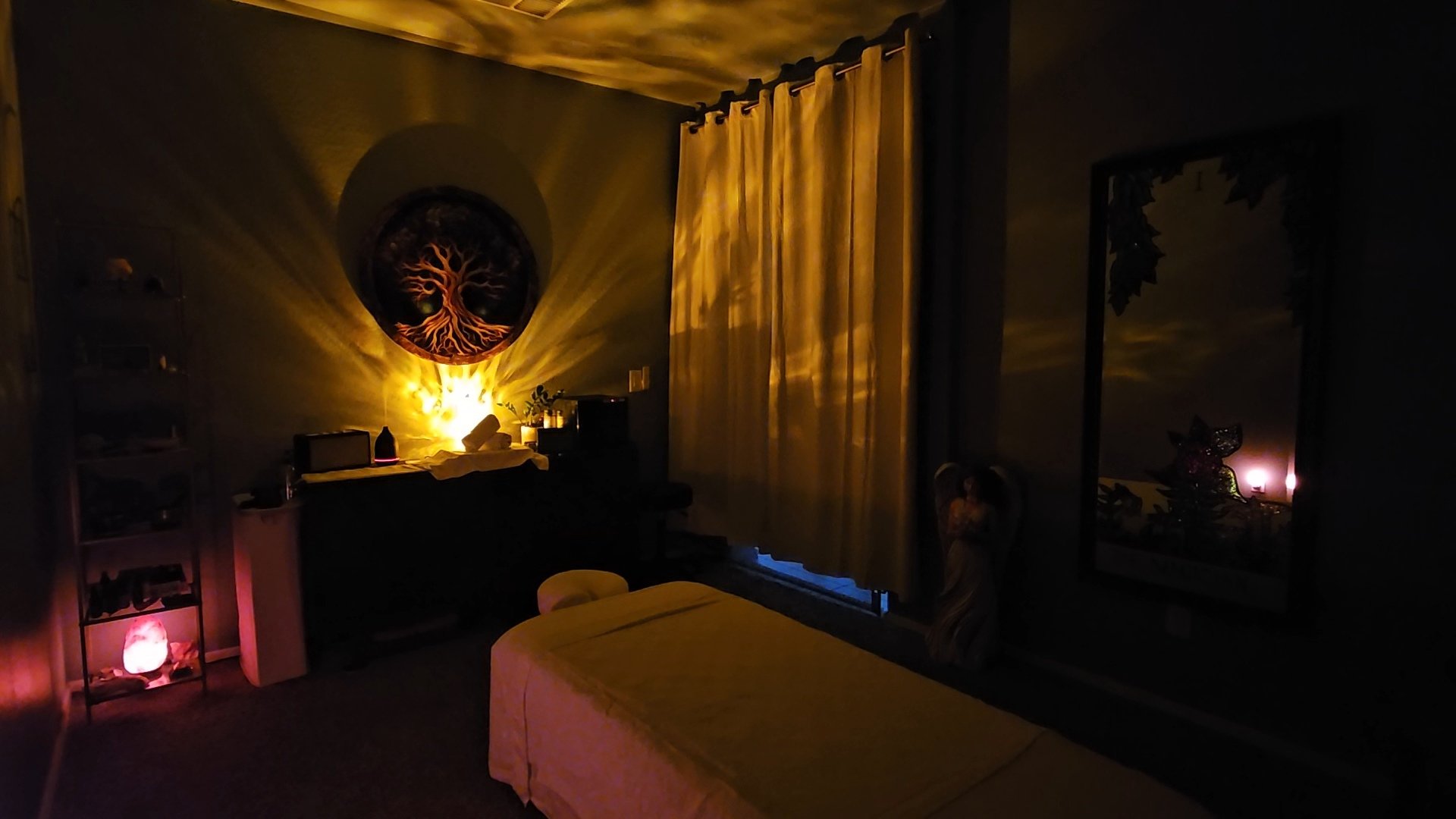 Dimly lit room with a massage table, curtains, a mirror, a large wall art of a tree, and ambient lighting