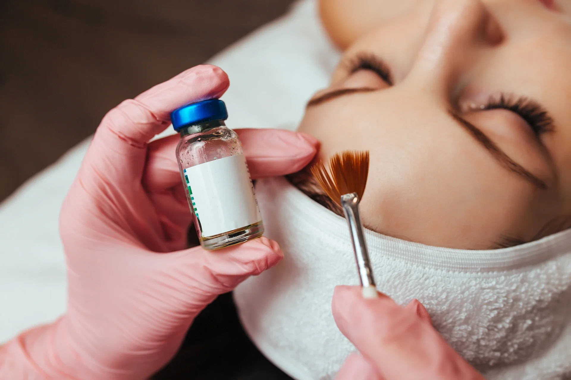 A person receiving a facial treatment, with a focus on the skincare procedure involving a small bottle and a brush applying liquid or serum to the cheek area.