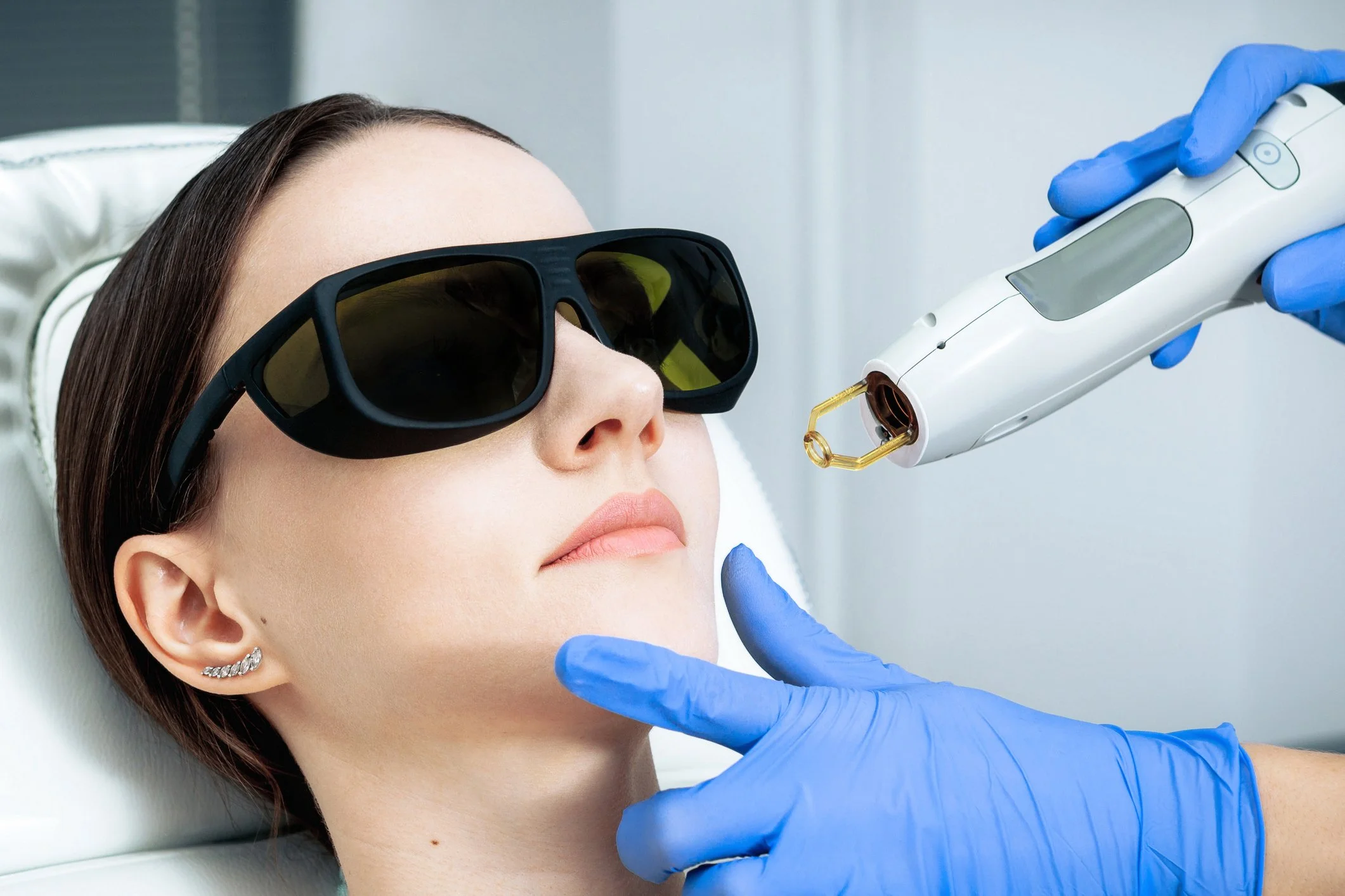A woman with brown hair wearing black sunglasses is lying down while a person wearing blue gloves uses a medical device near her face: skin lasers, skin regeneration, Cuterra Genesis. Triggering collagen and elastin production naturally.