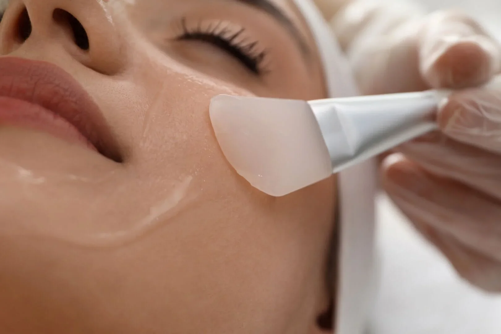 A person receiving a facial treatment with a spatula applying a skincare mask on their cheek.