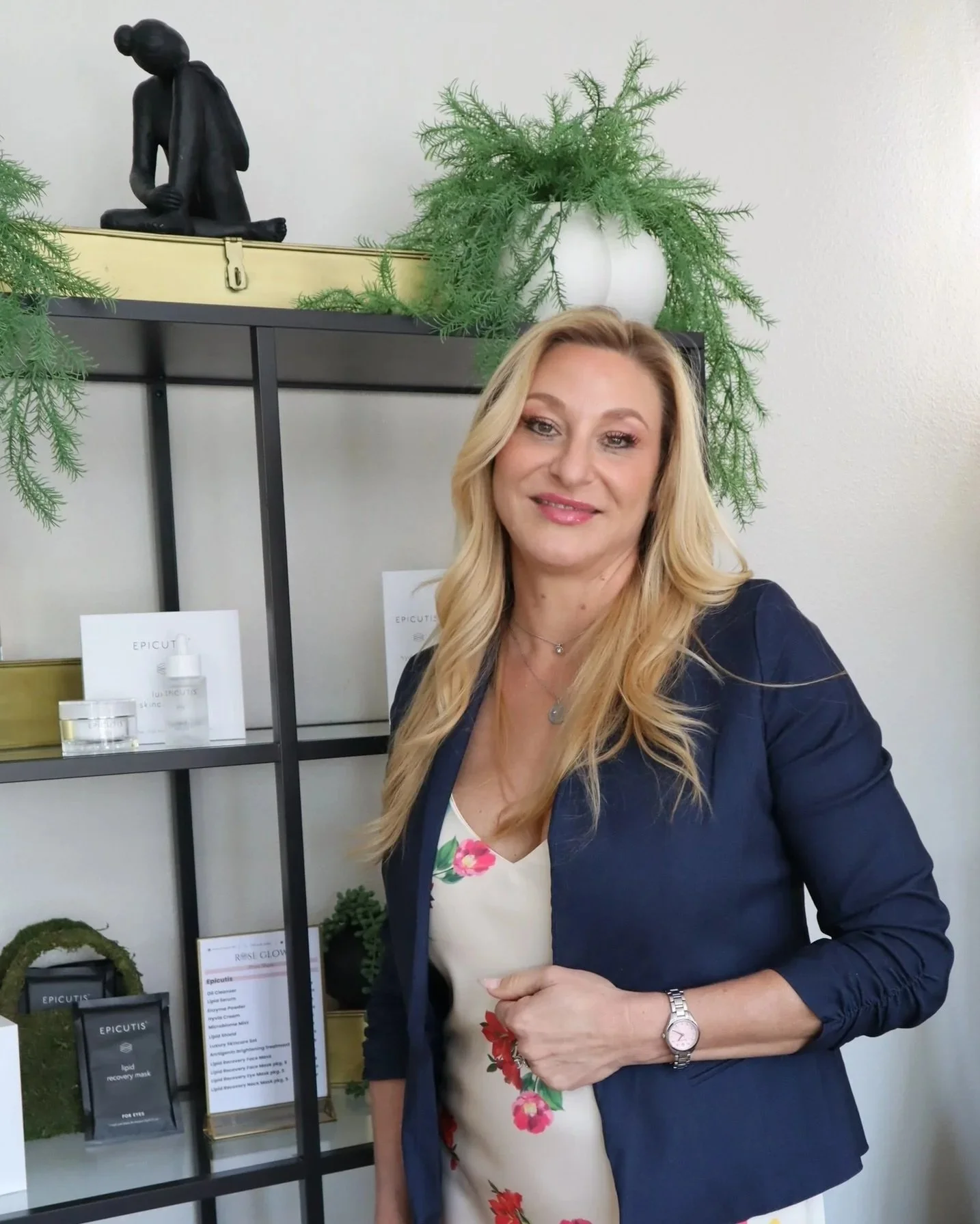 The owner and regishtered nurse with long blonde hair wearing a floral dress and navy blazer standing in front of a display shelf with skincare products and green plants, with a statue and white wall behind her.