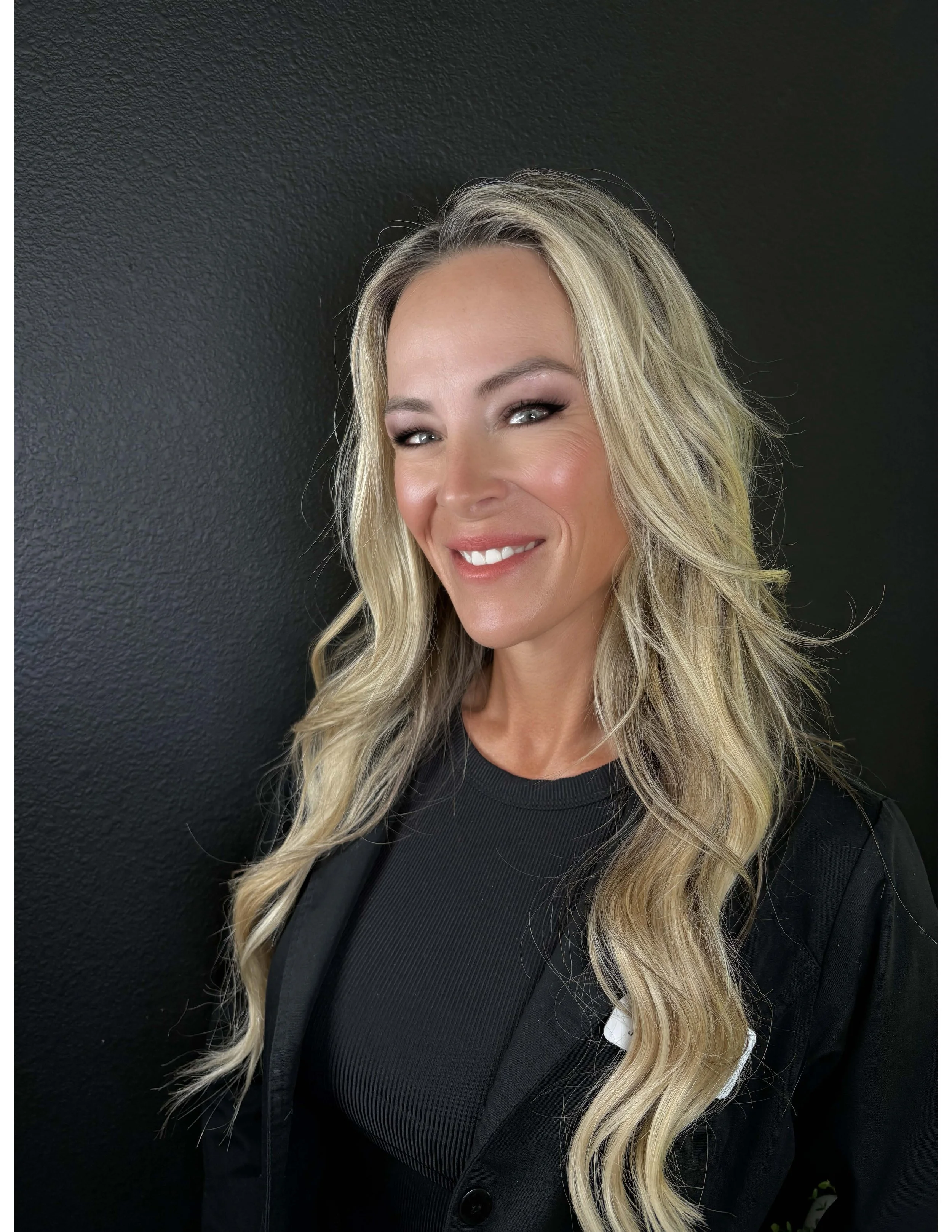 Allison Layton, an advanced laser technician, smiling woman with long blonde hair, wearing a black top and blazer, standing in front of a dark textured wall.